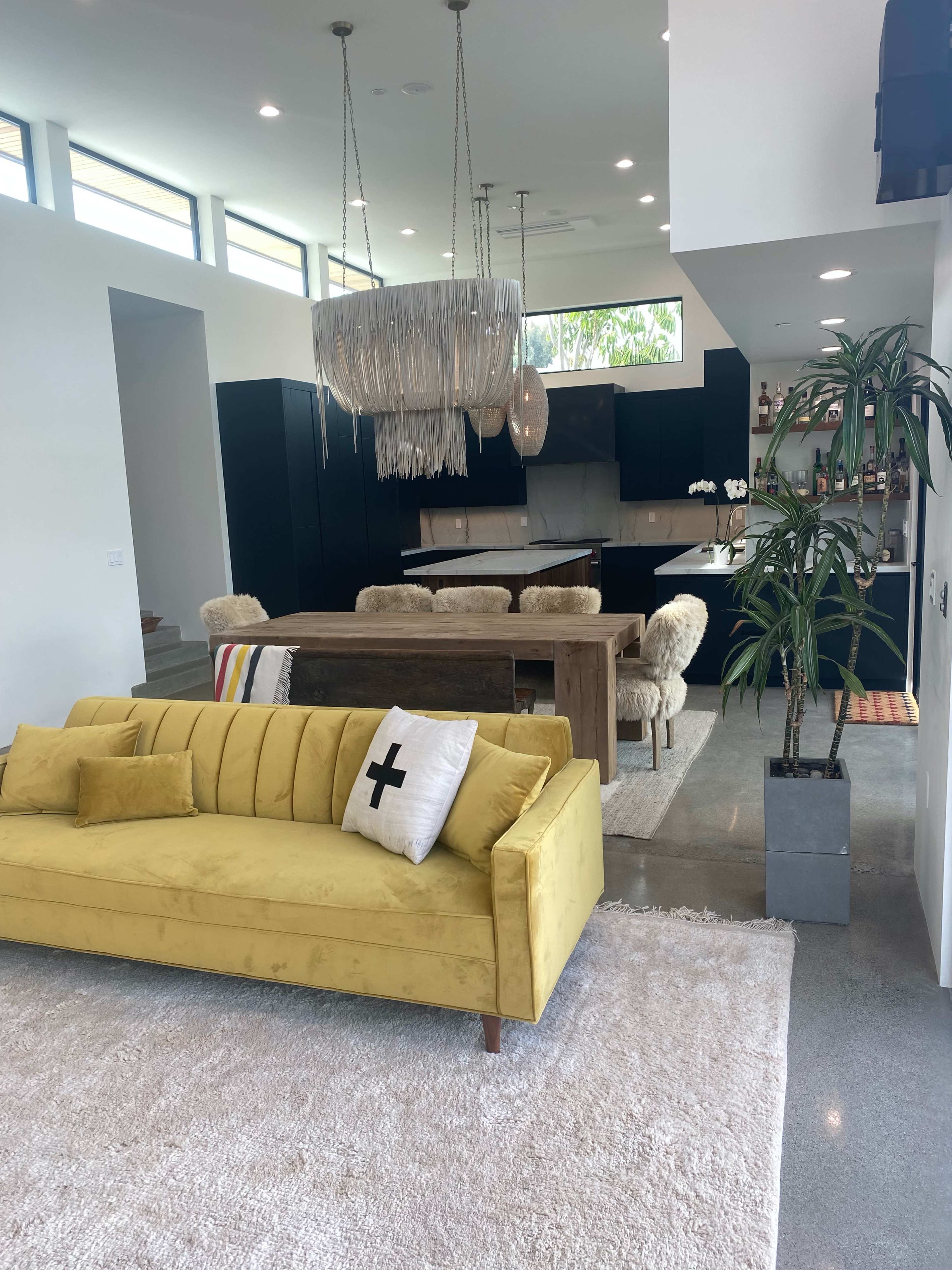 A modern living space featuring a yellow sofa in the foreground, a wooden dining table with fur-covered chairs in the background, and large windows near the ceiling.