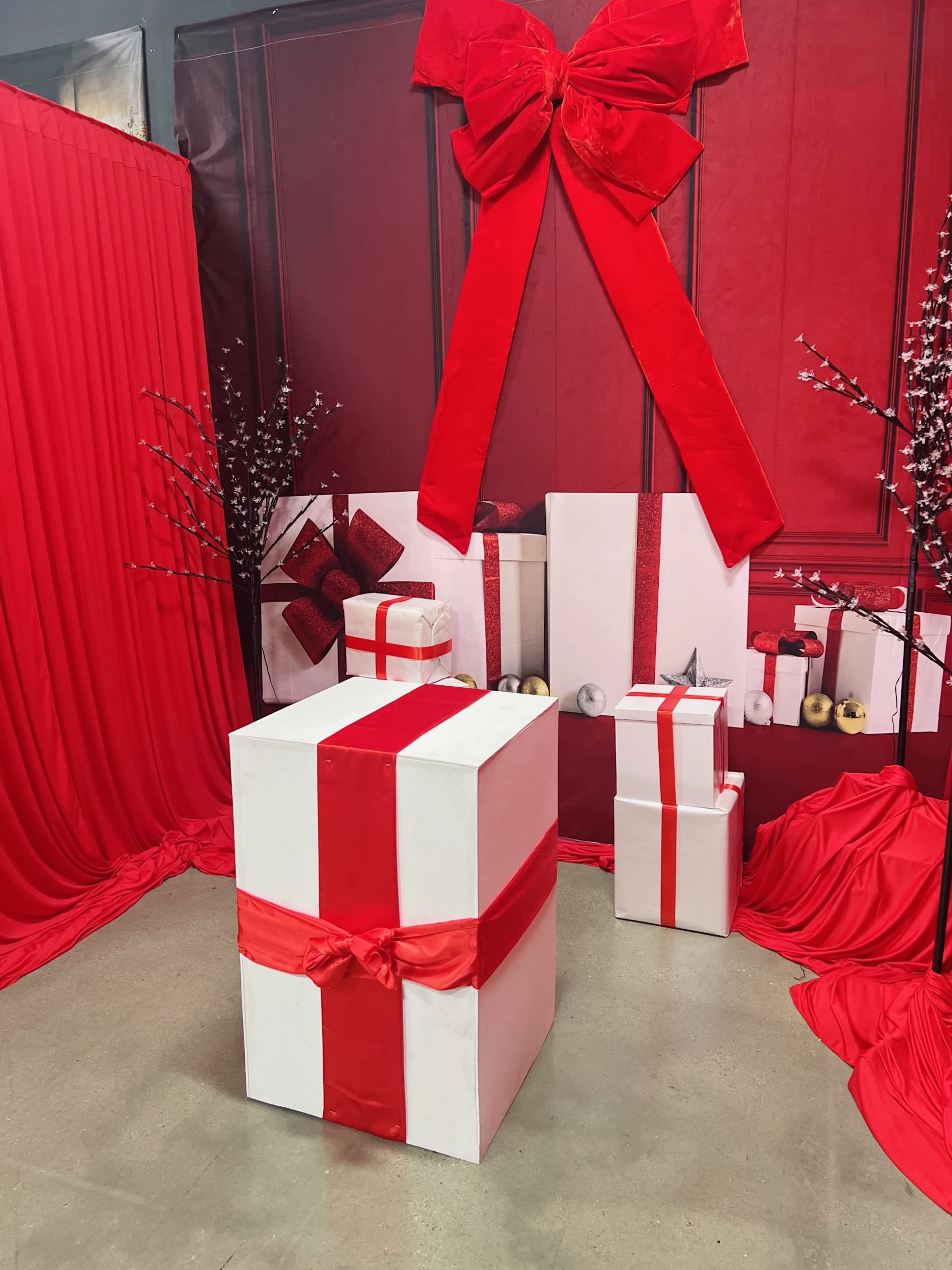 The scene features several large gift boxes wrapped in white paper with red ribbons, set against a backdrop of red drapery and decorations.