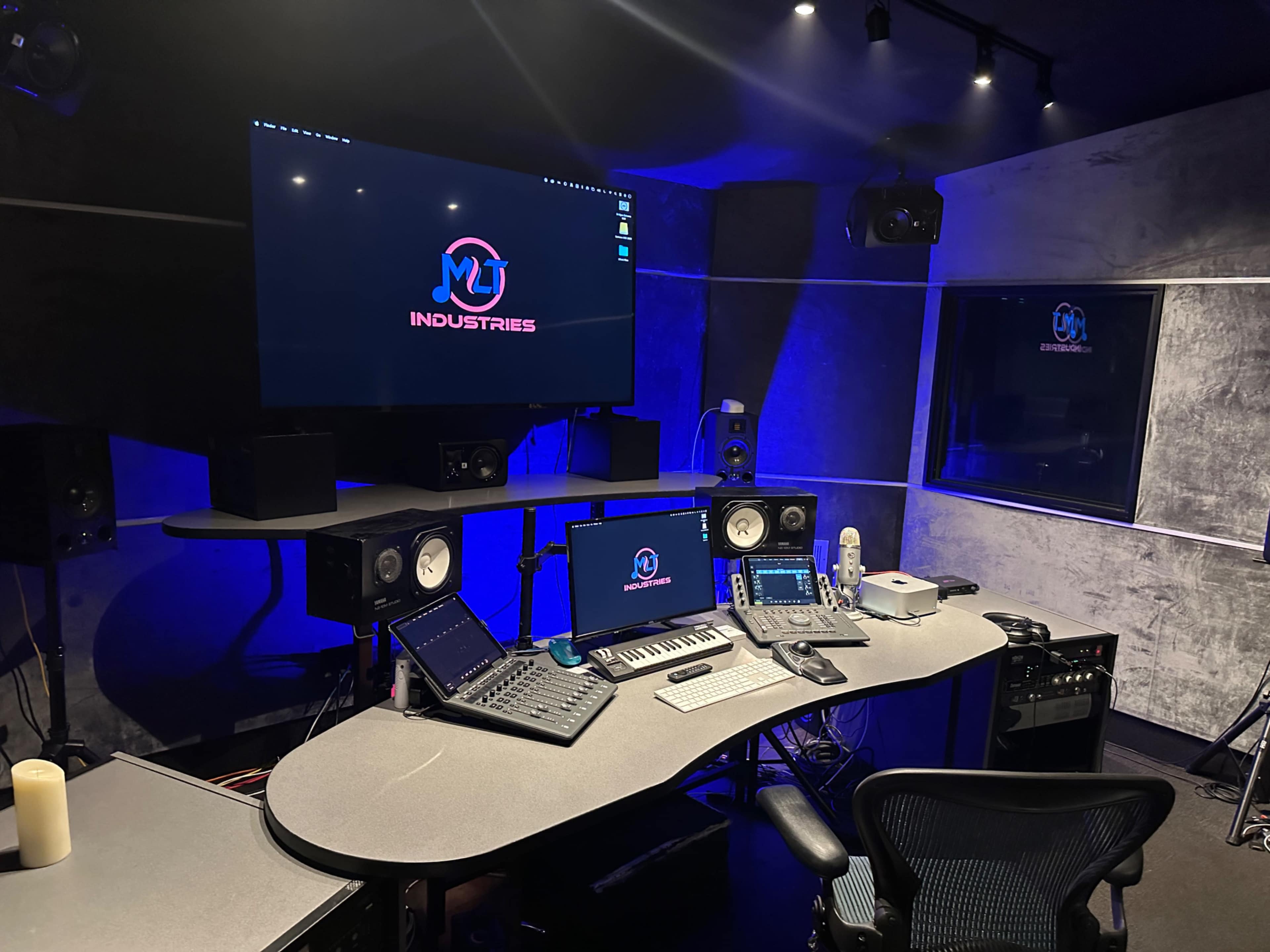 The image shows a modern music studio with a large display screen, mixing console, monitors, and audio equipment in a dimly lit environment.