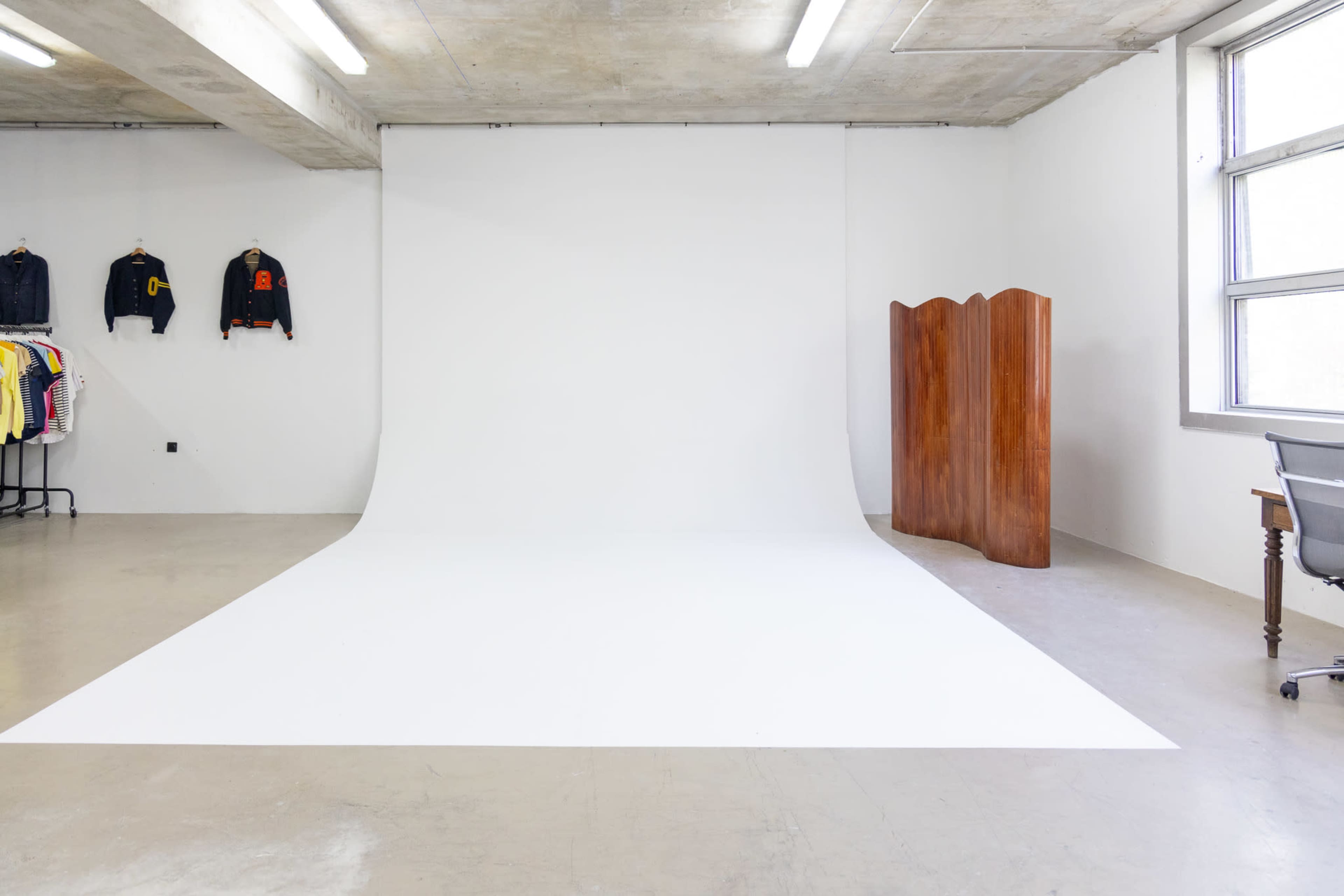 The image shows an empty photo studio with a white backdrop, a wooden room divider, and a desk with a chair.