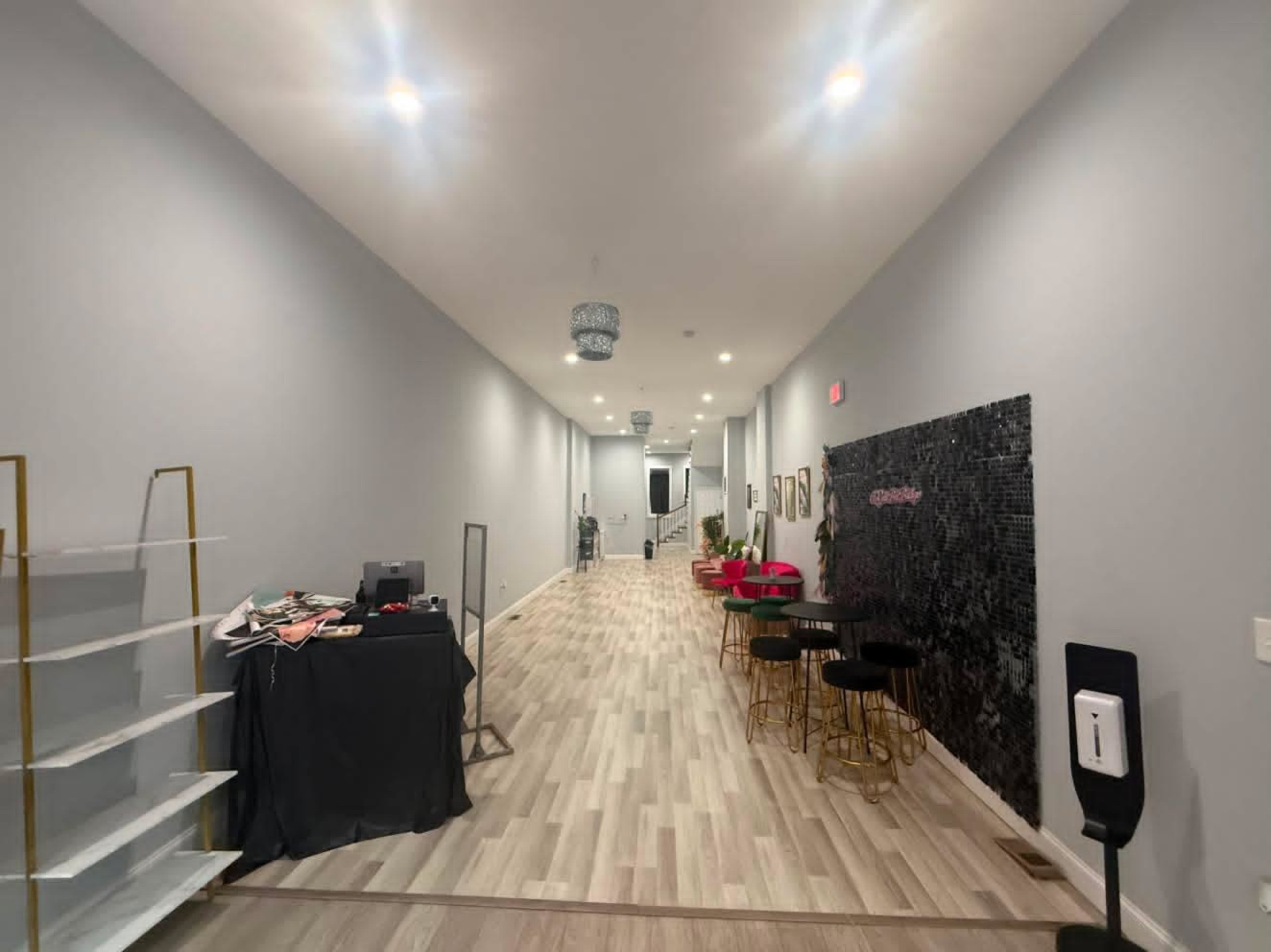 The image depicts a long, well-lit hallway with a sleek, modern design featuring light-colored walls, wooden flooring, and decorative elements including a black mosaic wall and a small seating area.