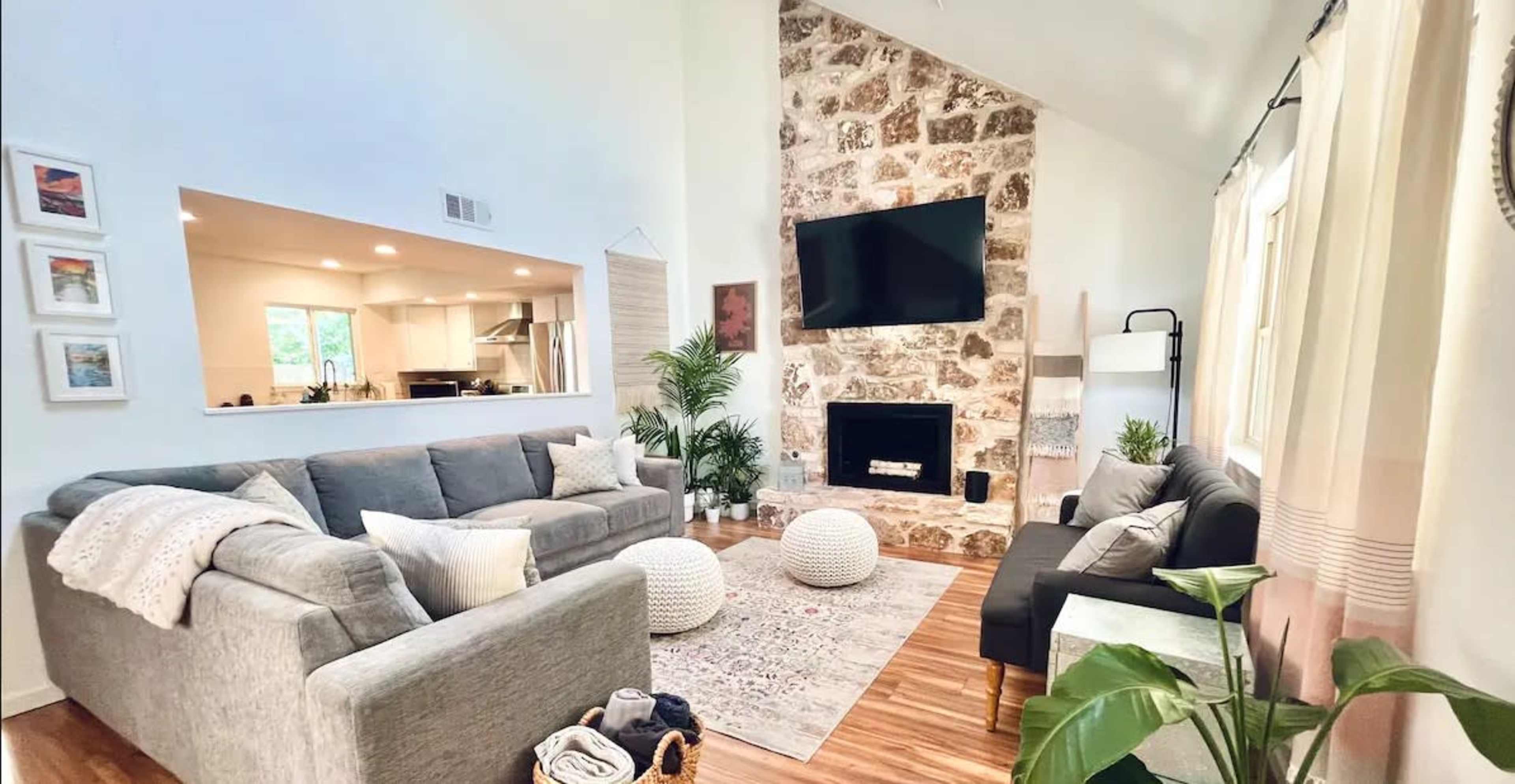 The living room features a stone fireplace, two distinct seating areas with gray sofas, and light-colored walls adorned with plants and decorative elements.