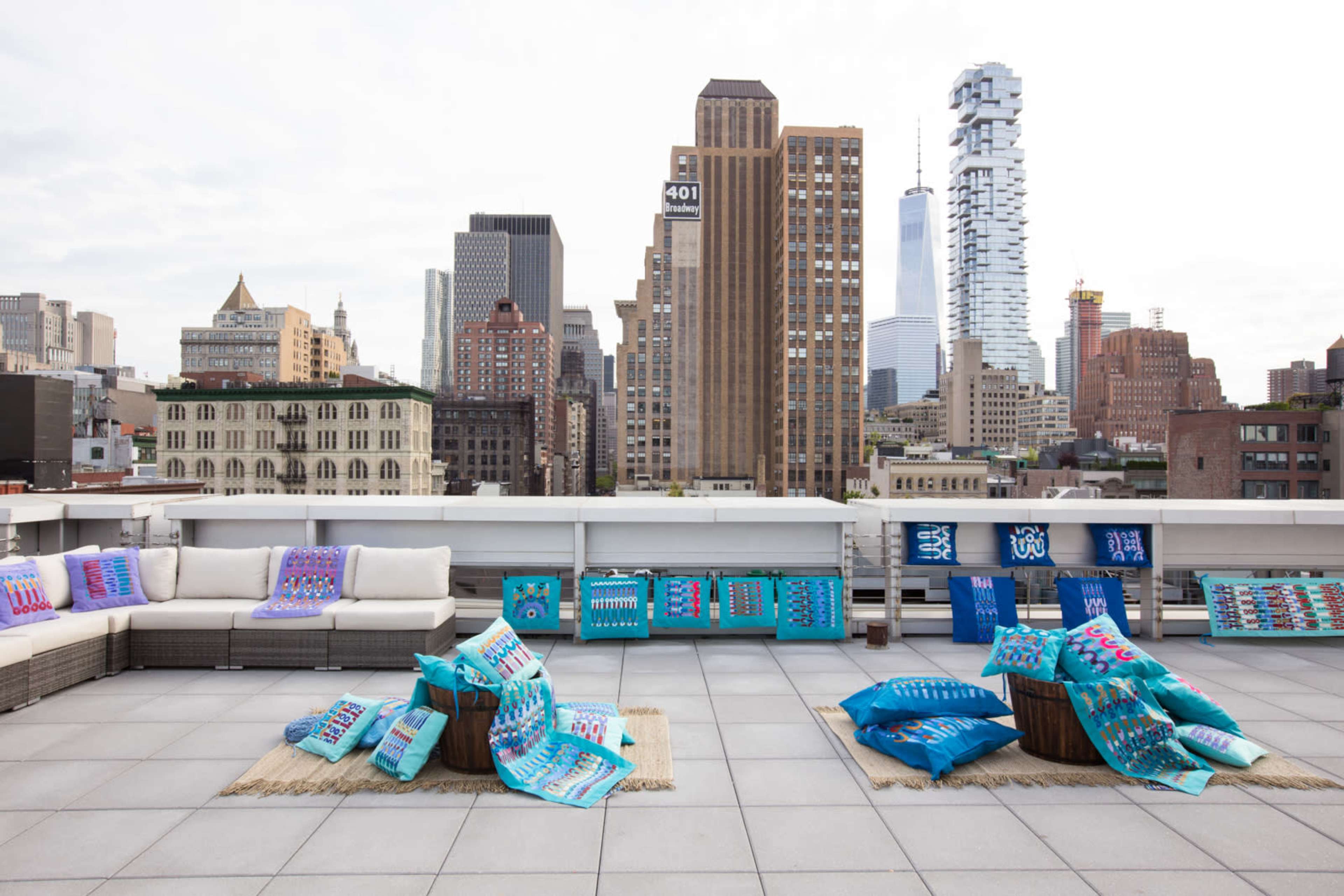 A rooftop space features colorful cushions and throws arranged on the ground, with a view of tall buildings and the skyline in the background.