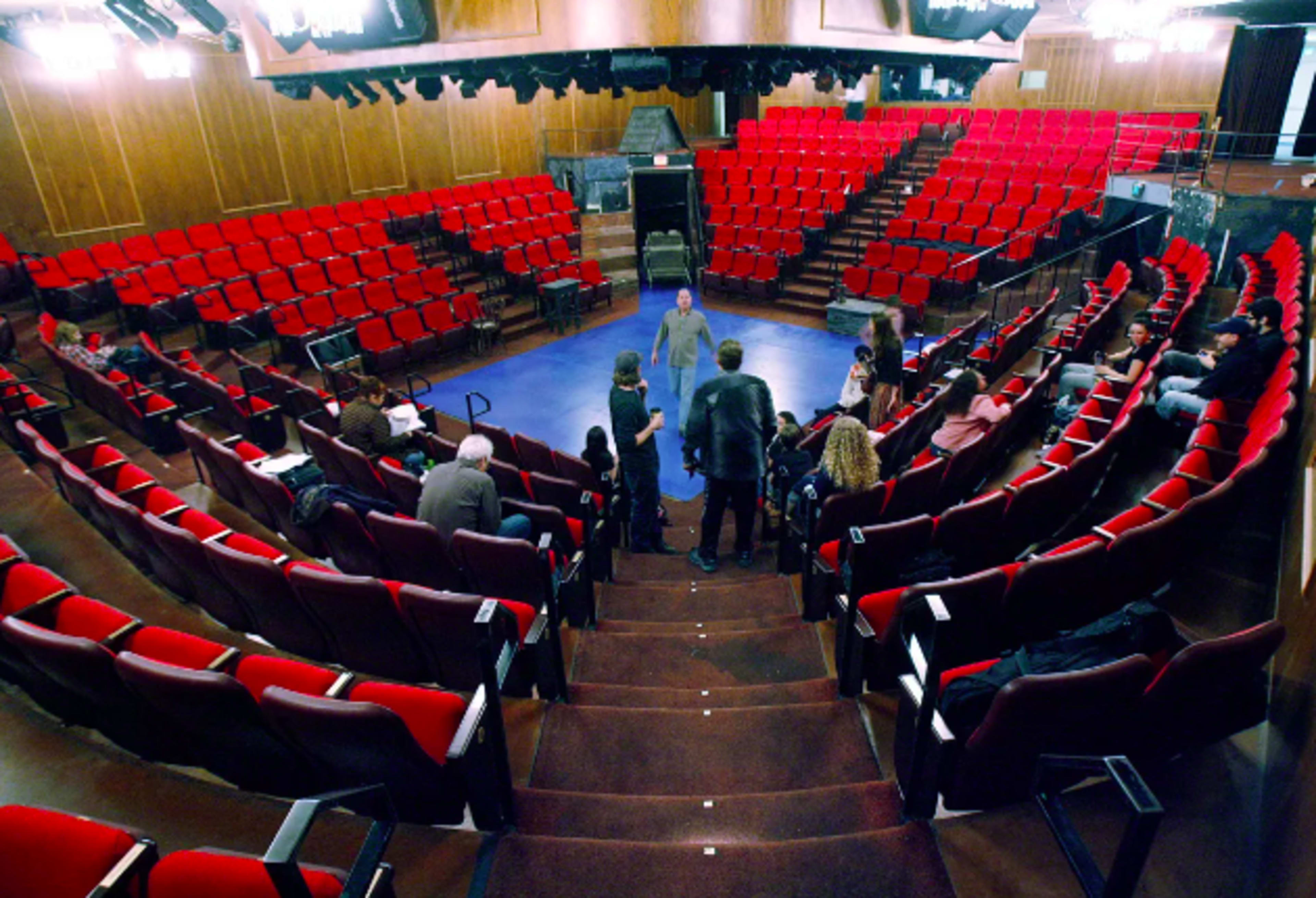 The image shows a theater with red seating arranged in a semi-circle around a central performance area, where several people are gathered on stage.
