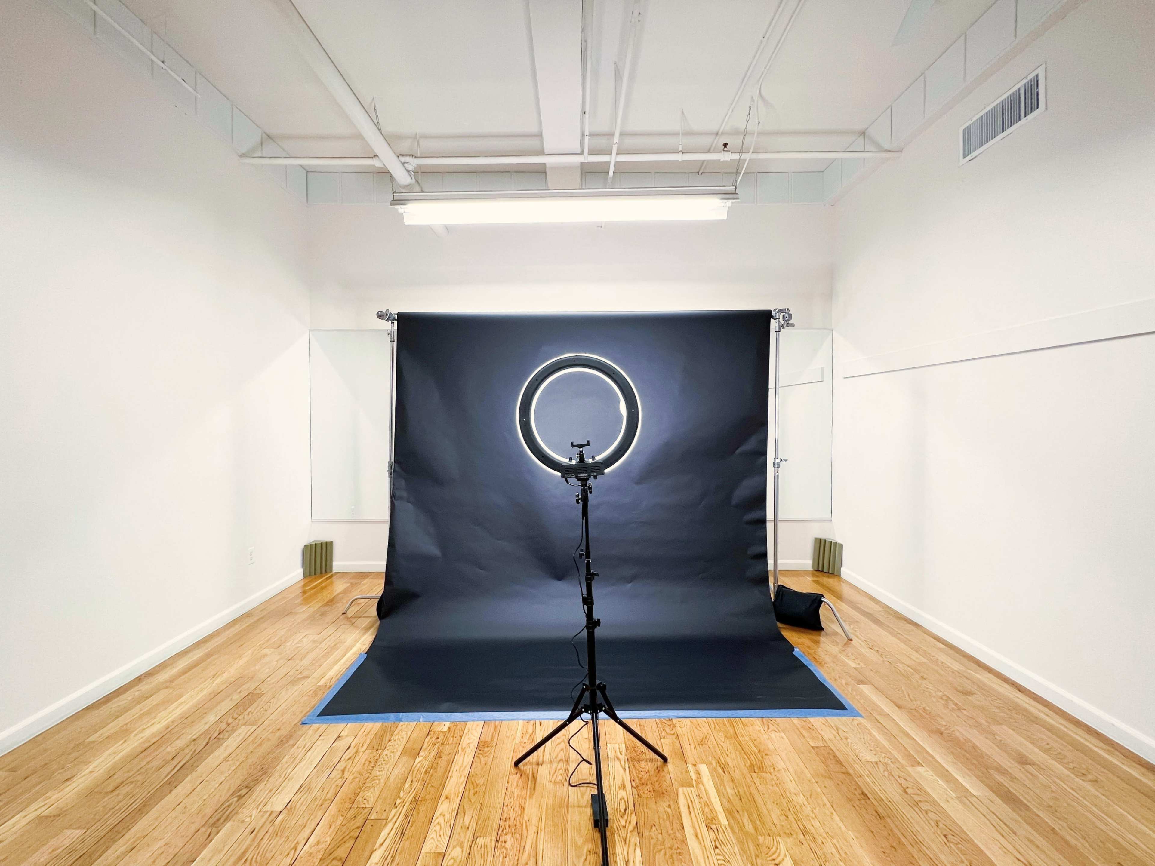 A studio space features a dark backdrop set up with a ring light on a tripod, surrounded by wooden flooring.