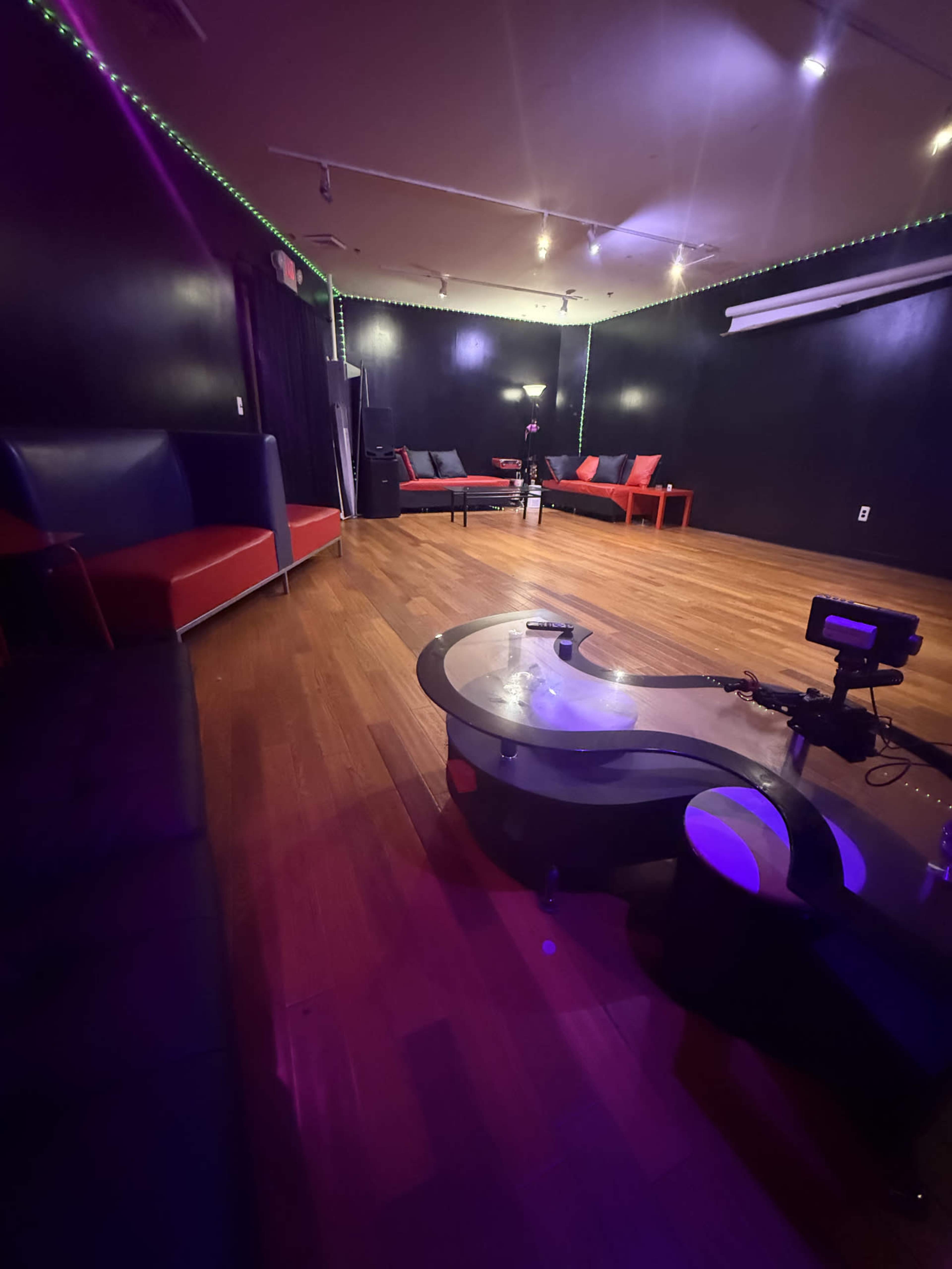 The image shows a dimly lit room with hardwood floors, featuring a few red and black couches and a central table.