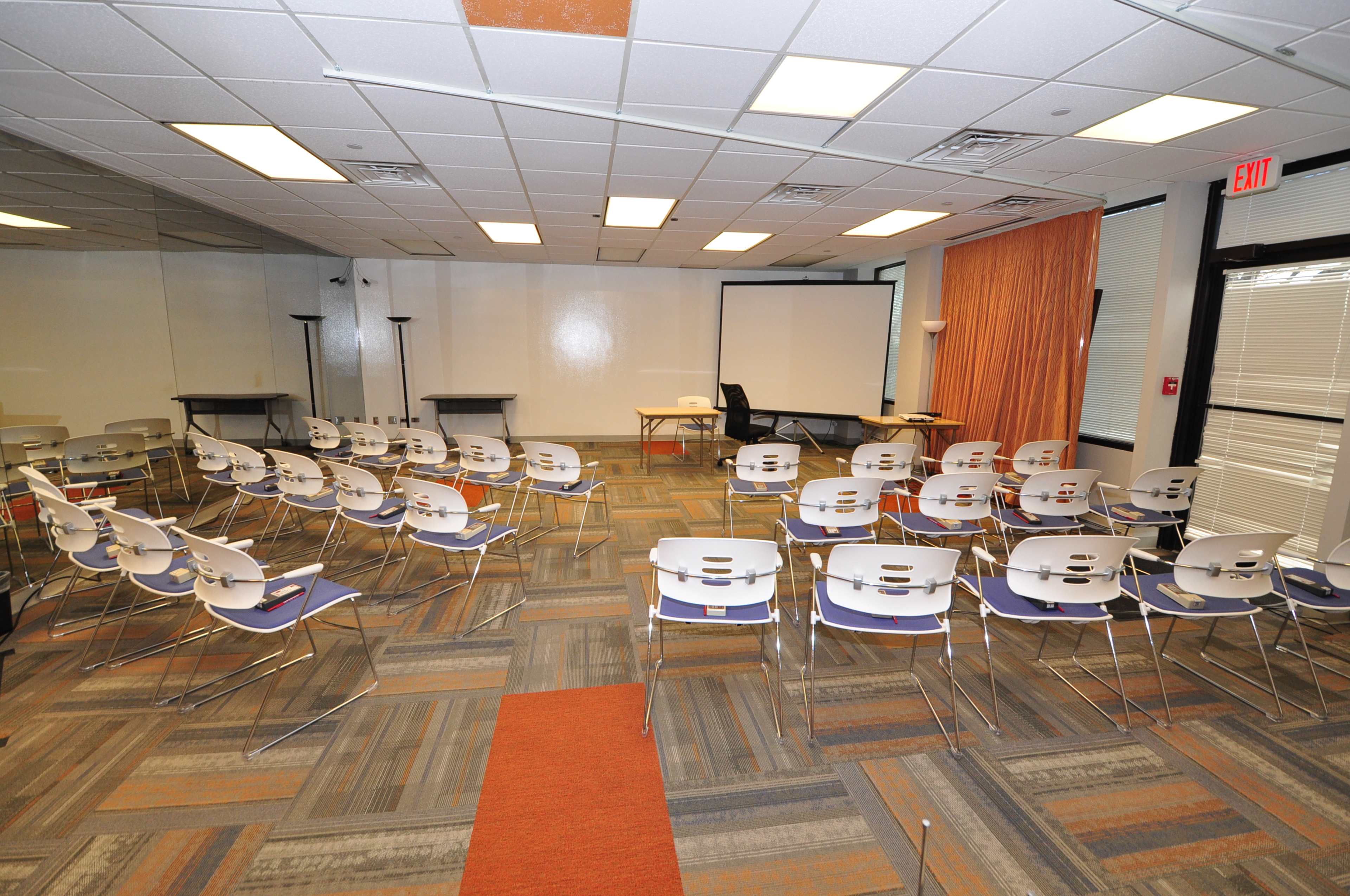 The image shows a classroom setup with rows of chairs facing a presentation screen and two tables in the background.