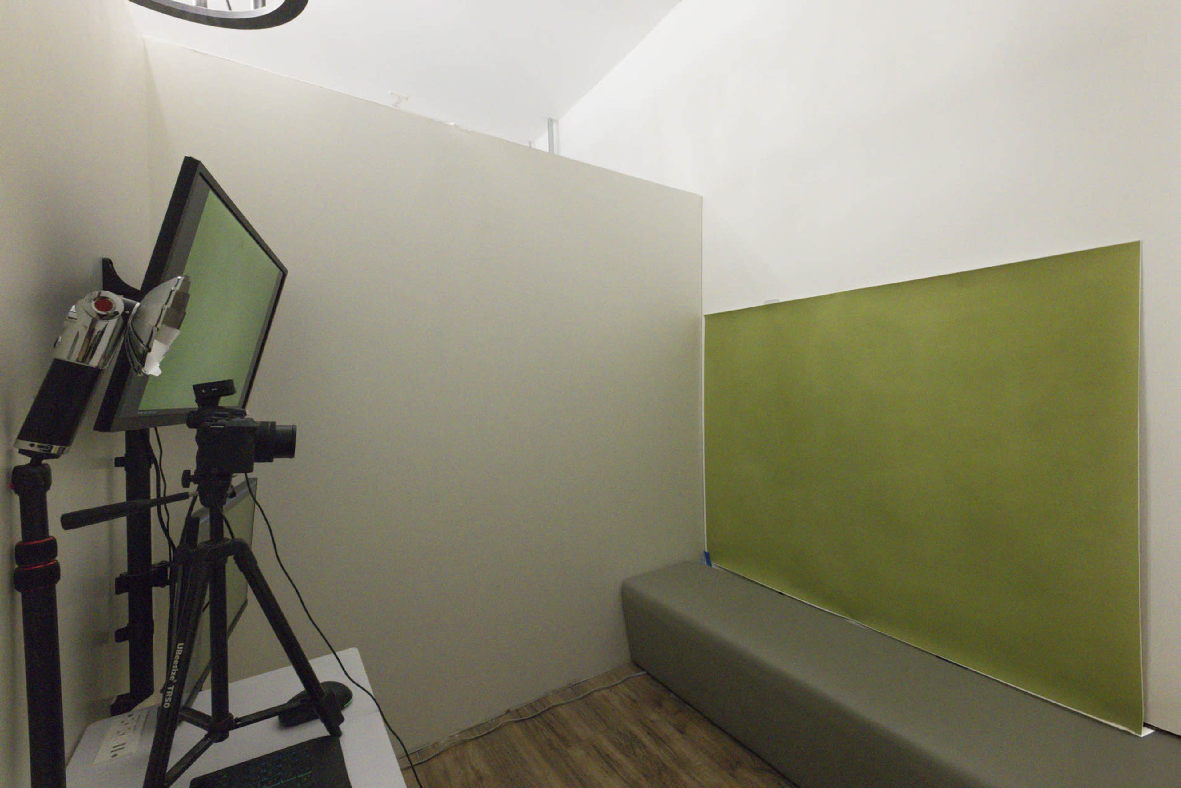 The image shows a small room equipped with a camera on a tripod, a monitor, a green wall section, and a gray bench.