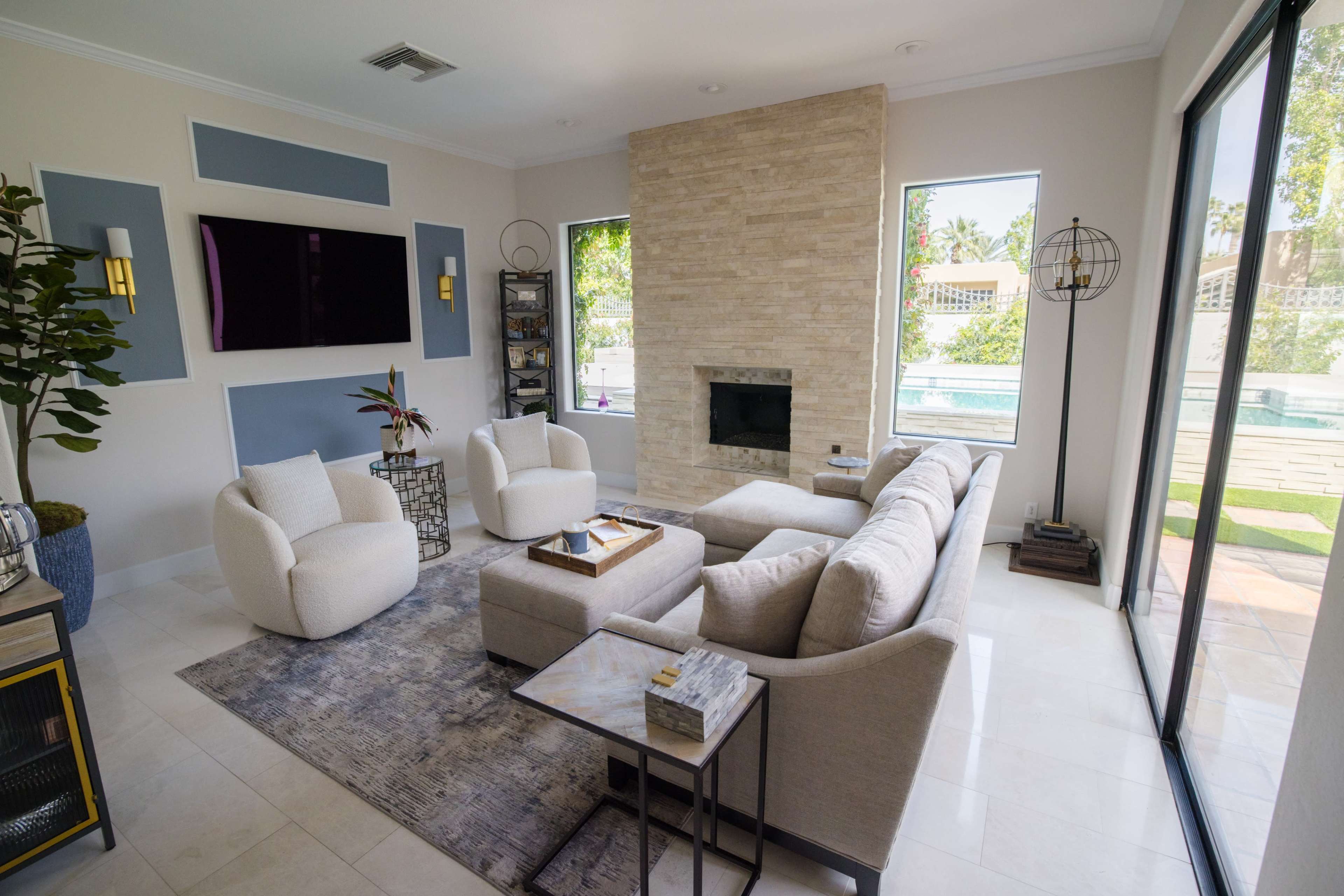 A modern living room featuring a sectional sofa, a coffee table, a stone fireplace, and large windows overlooking a pool area.