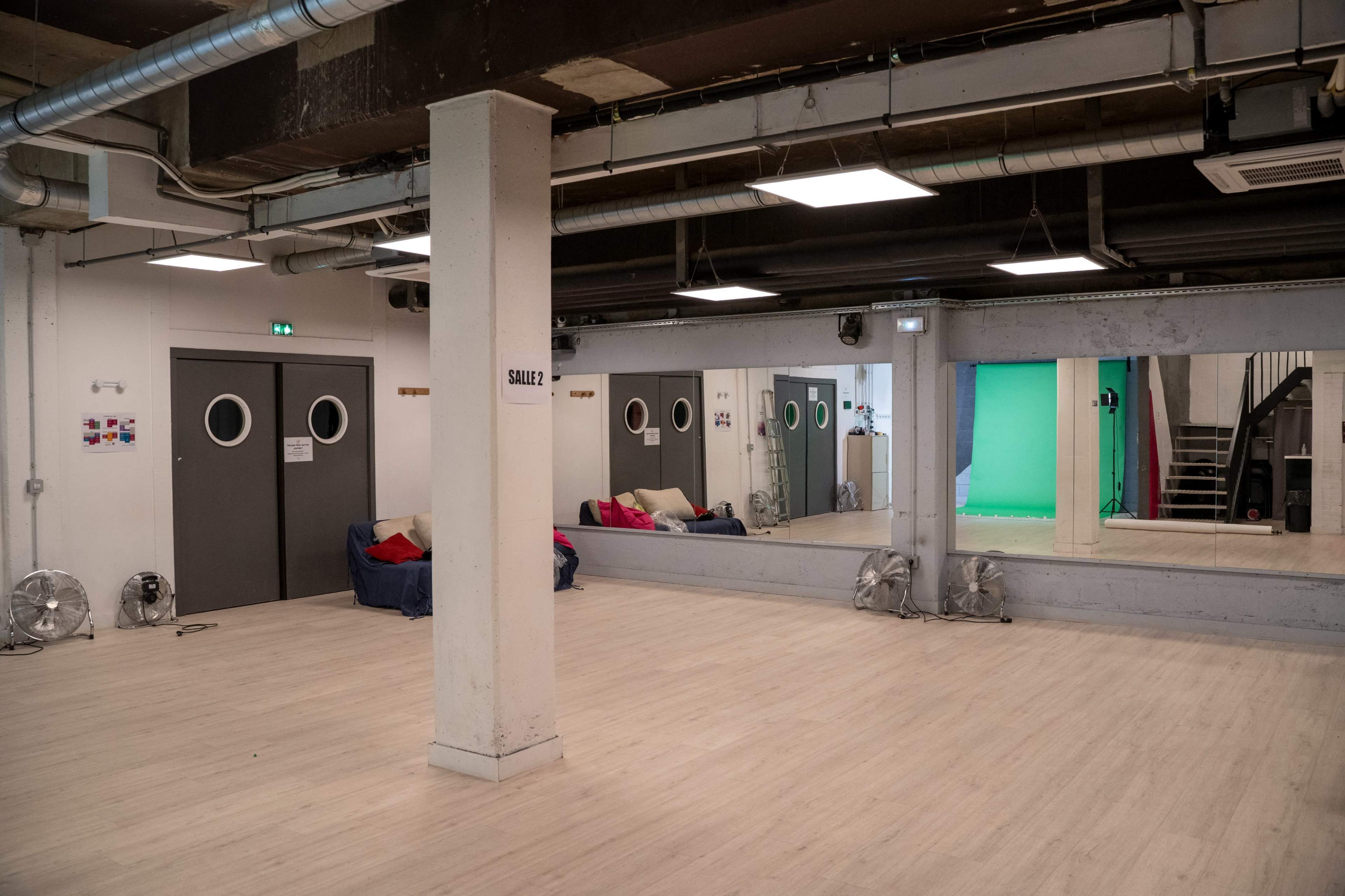 A spacious room with mirrors, a staircase, and a green screen area, featuring seating along one wall.