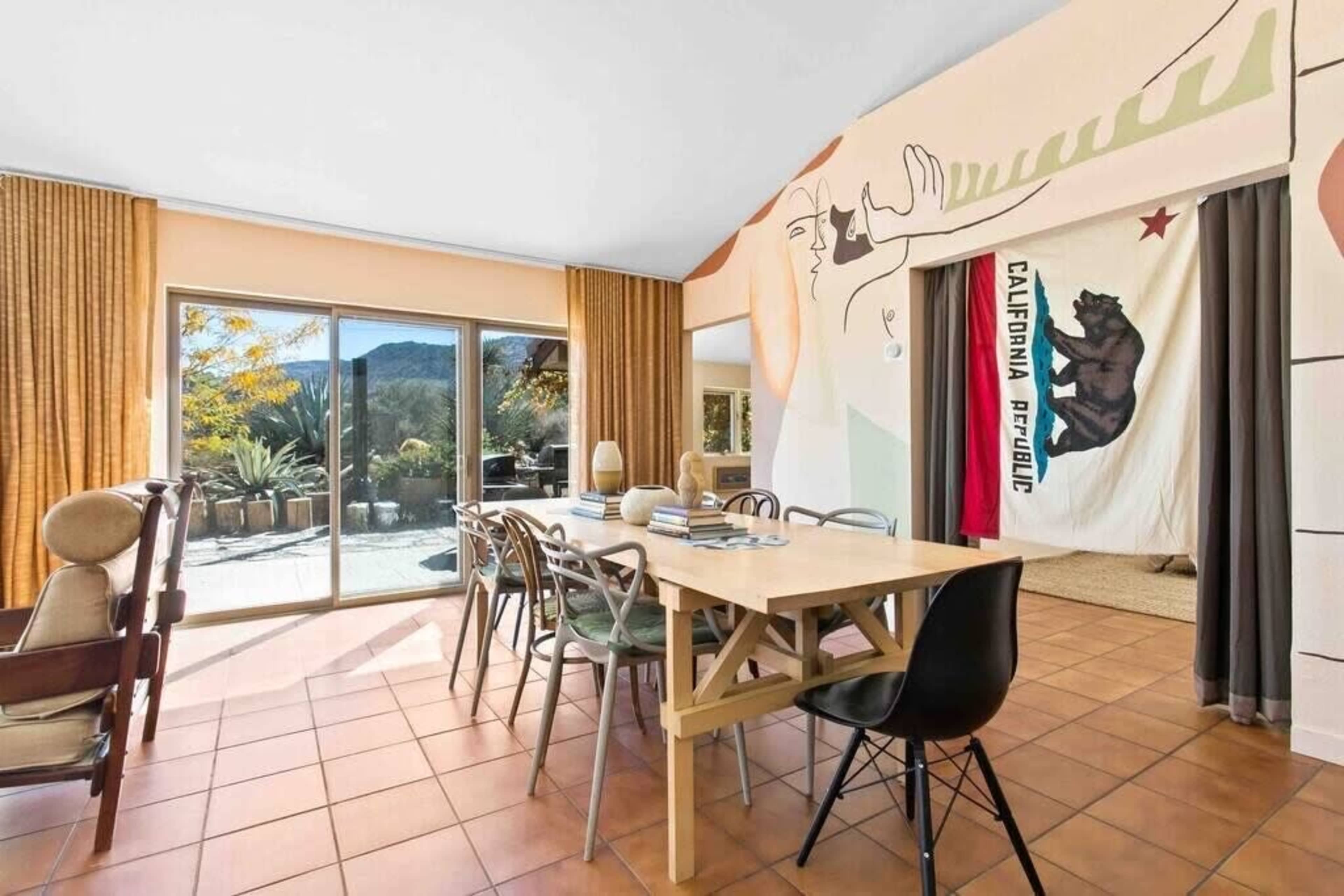 A modern dining area with a large table, a mix of dining chairs, and a wall mural, leading to a patio with outdoor views.