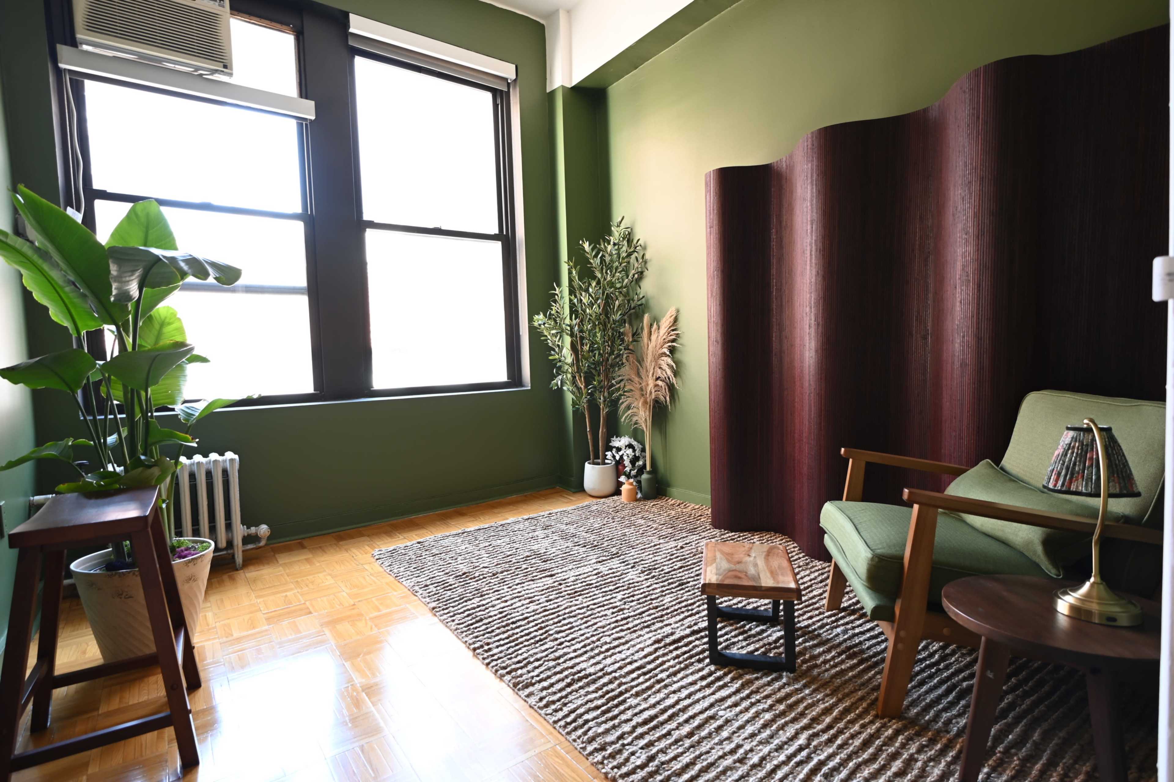 The image shows a minimalist room with green walls, a wooden screen, a green armchair, a coffee table, a plant, and large windows allowing natural light to enter.