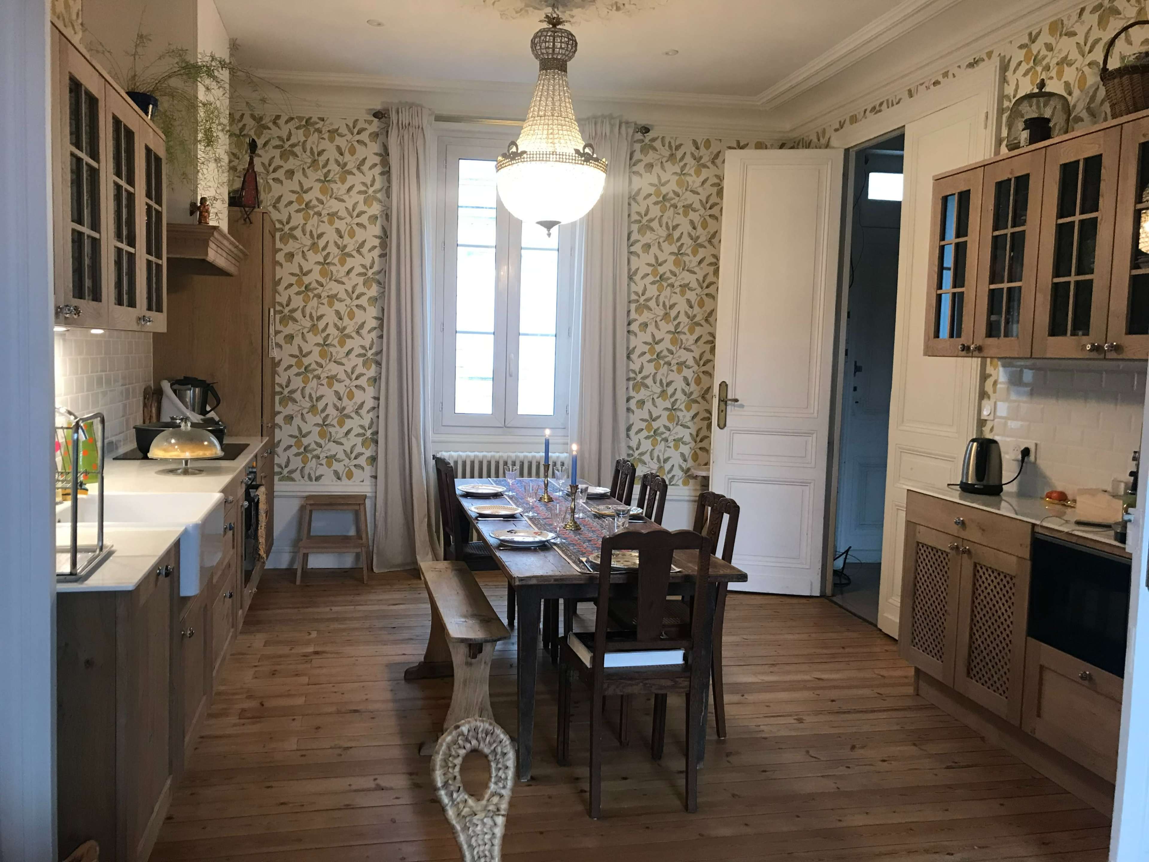 A light-filled kitchen features wooden cabinets, a dining table set for a meal, and floral wallpaper.