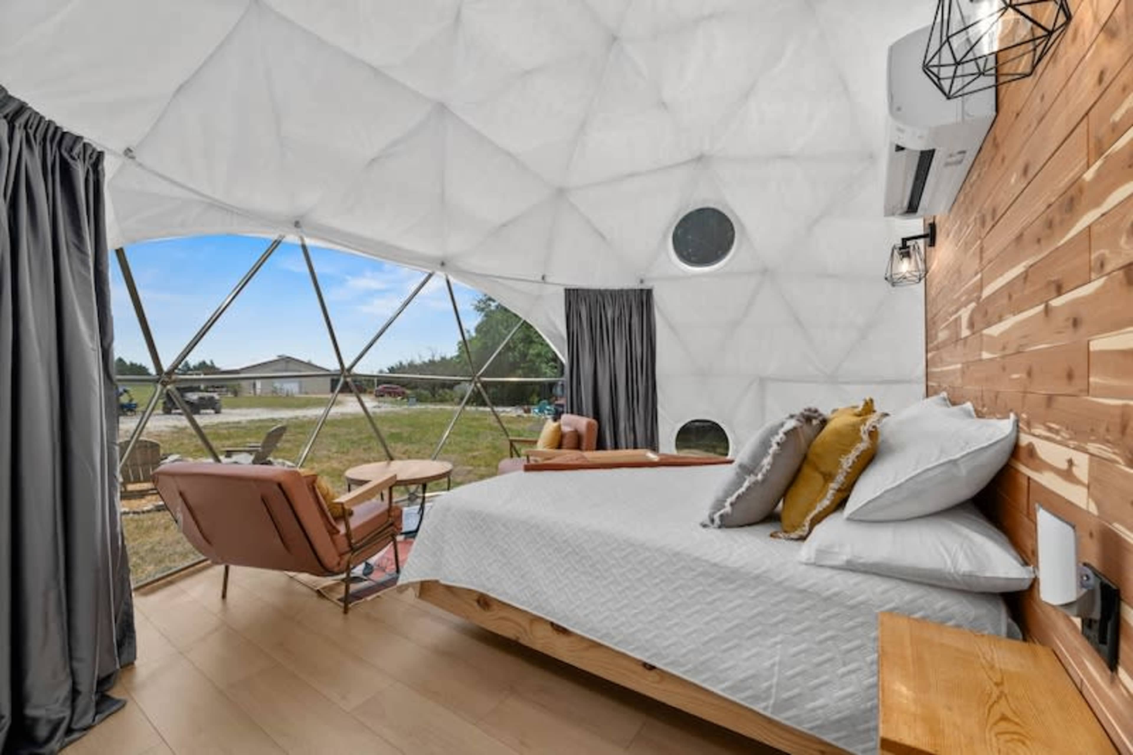 The interior of a geodesic dome features a wooden bed, two armchairs, and large triangular windows overlooking a grassy outdoor area.