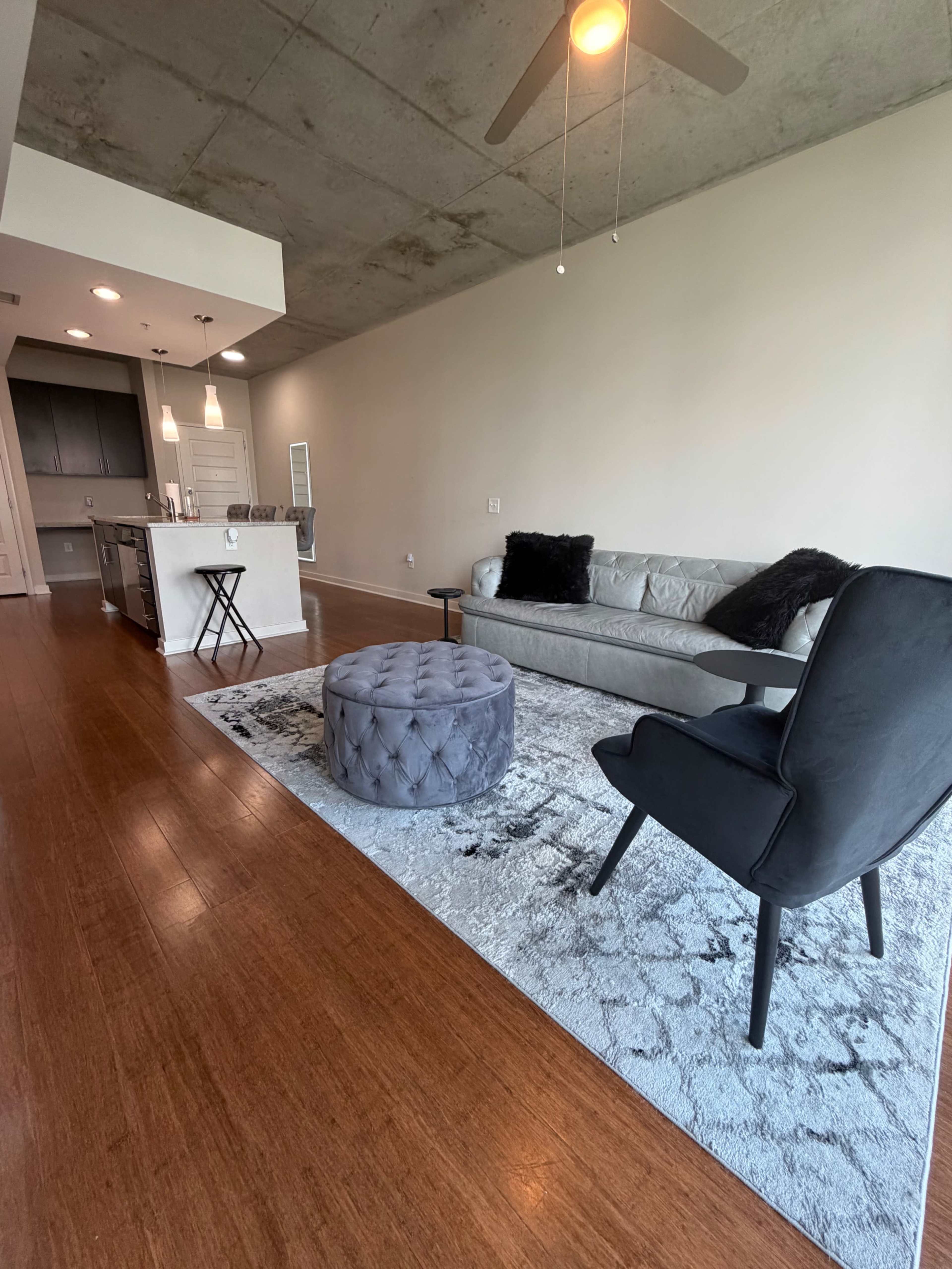 The image shows a modern living room with a gray sofa, a round tufted ottoman, and a black armchair, all arranged on a plush area rug.