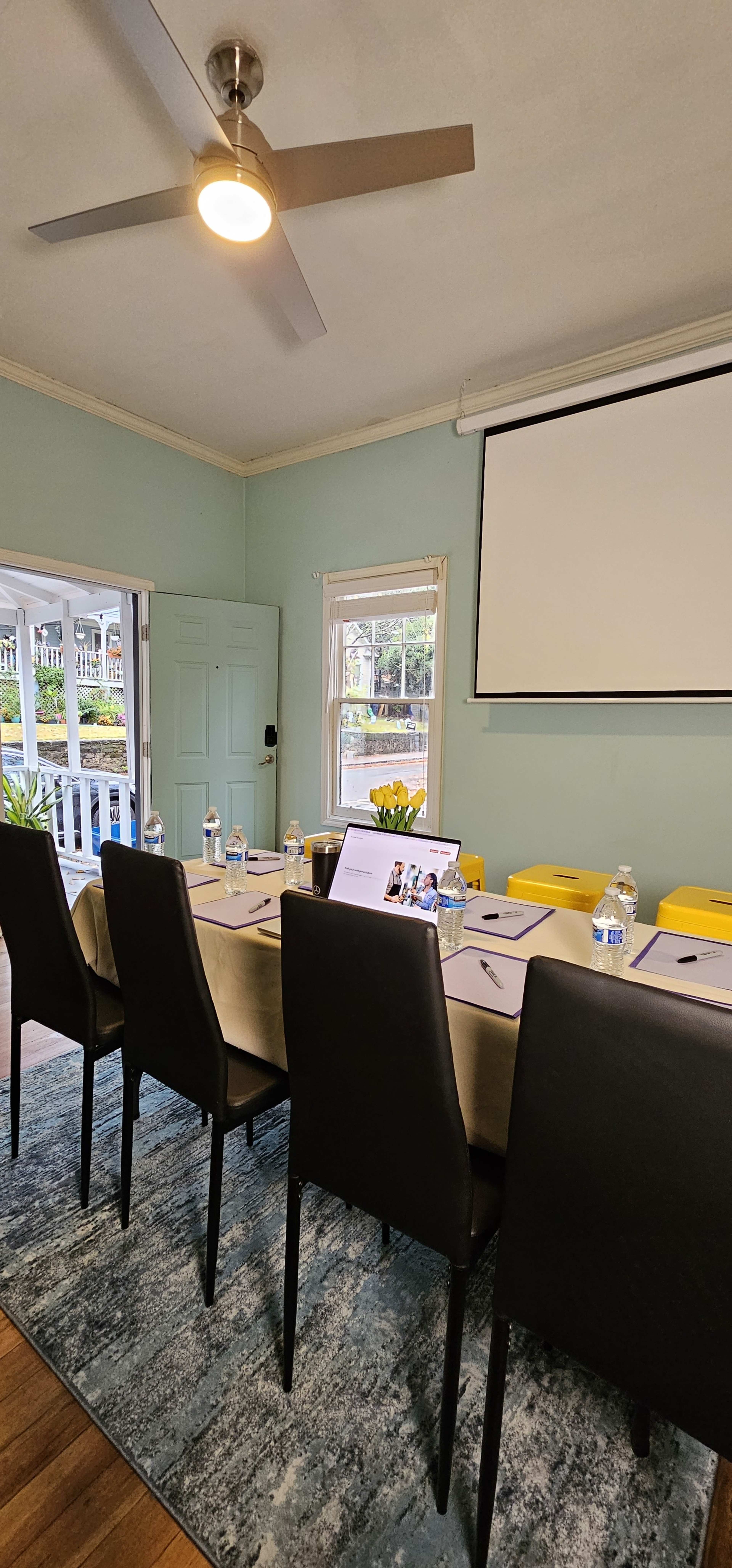 A conference room is set up with a long table, chairs, water bottles, and a projector screen, with natural light coming from a nearby window.