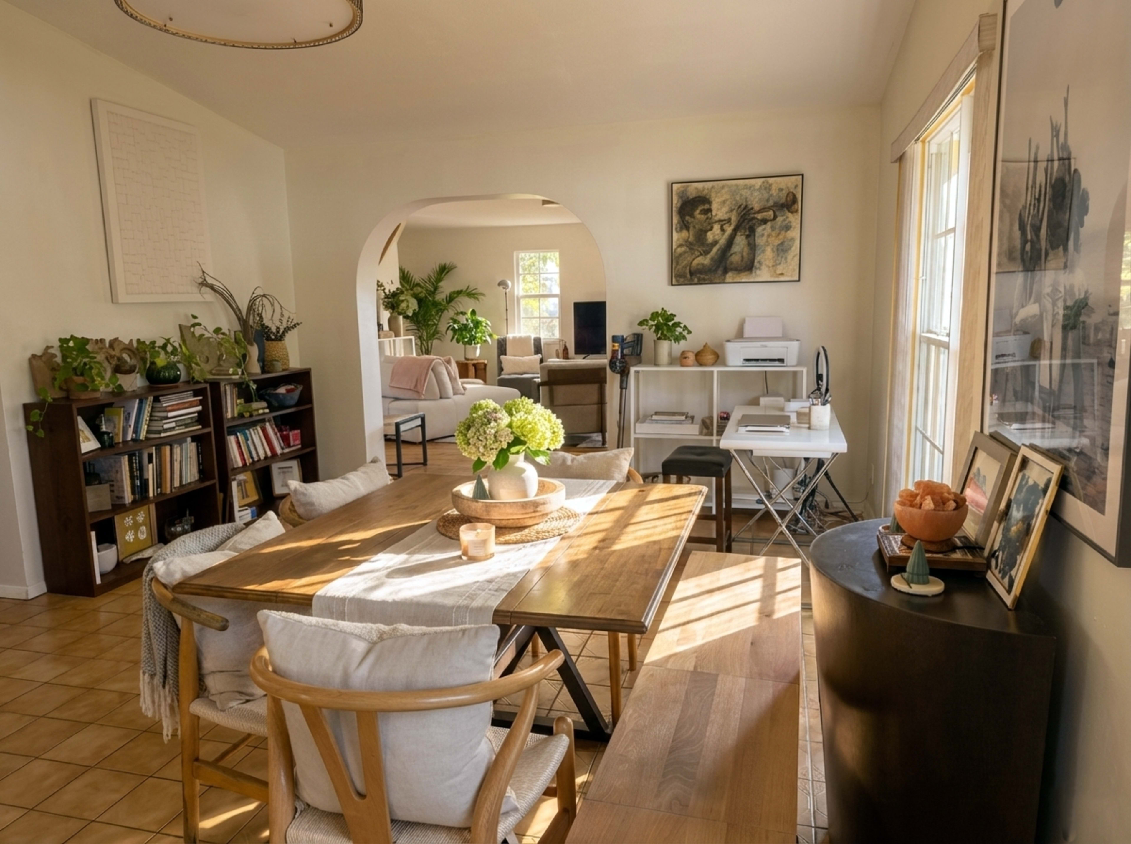 The image shows a well-lit dining area with a wooden table, surrounded by chairs, and divided from a living space by an archway, featuring various plants and furniture.