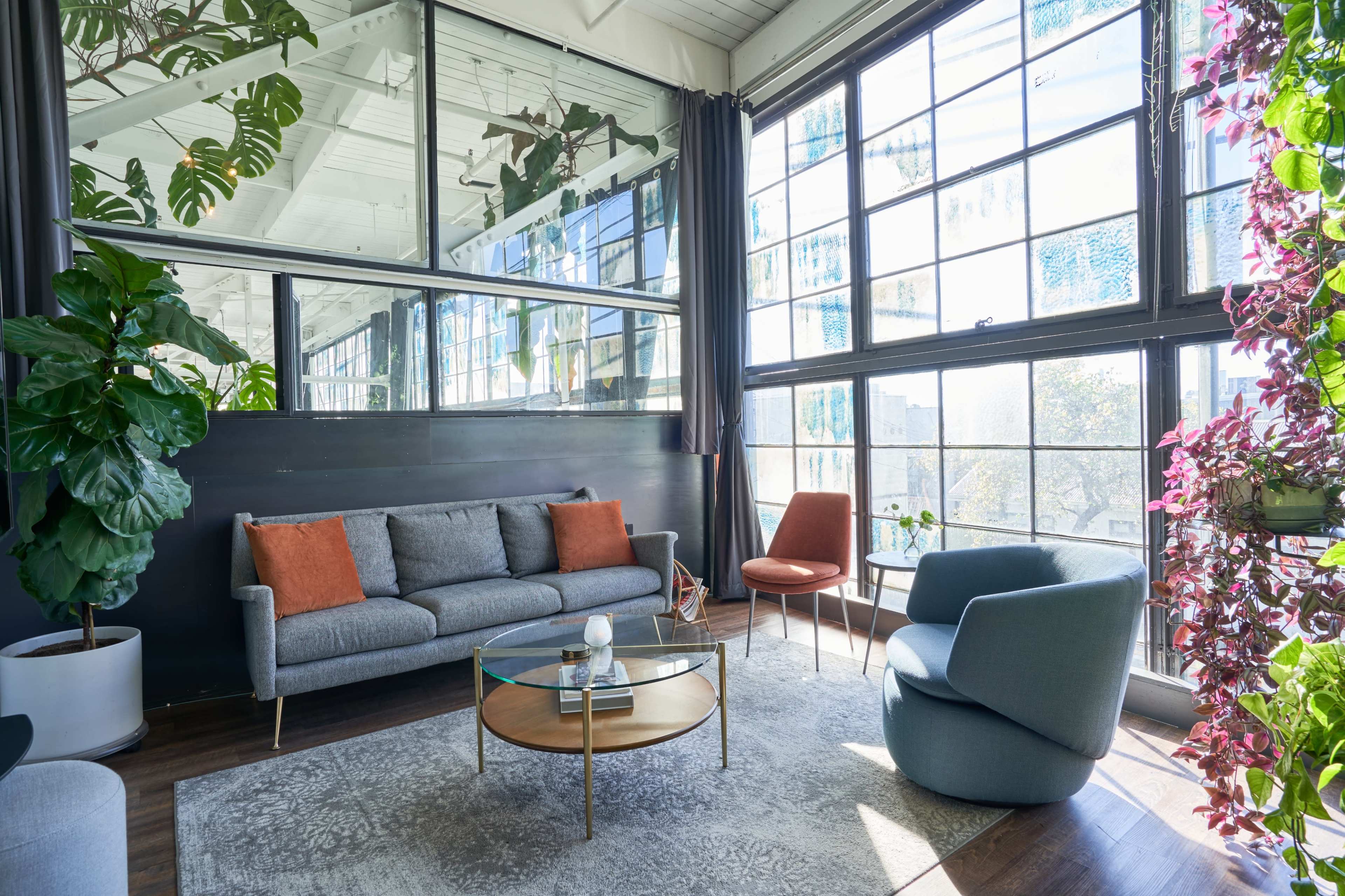 A modern living room features a gray sofa, an orange accent chair, a blue armchair, and large windows surrounded by greenery.