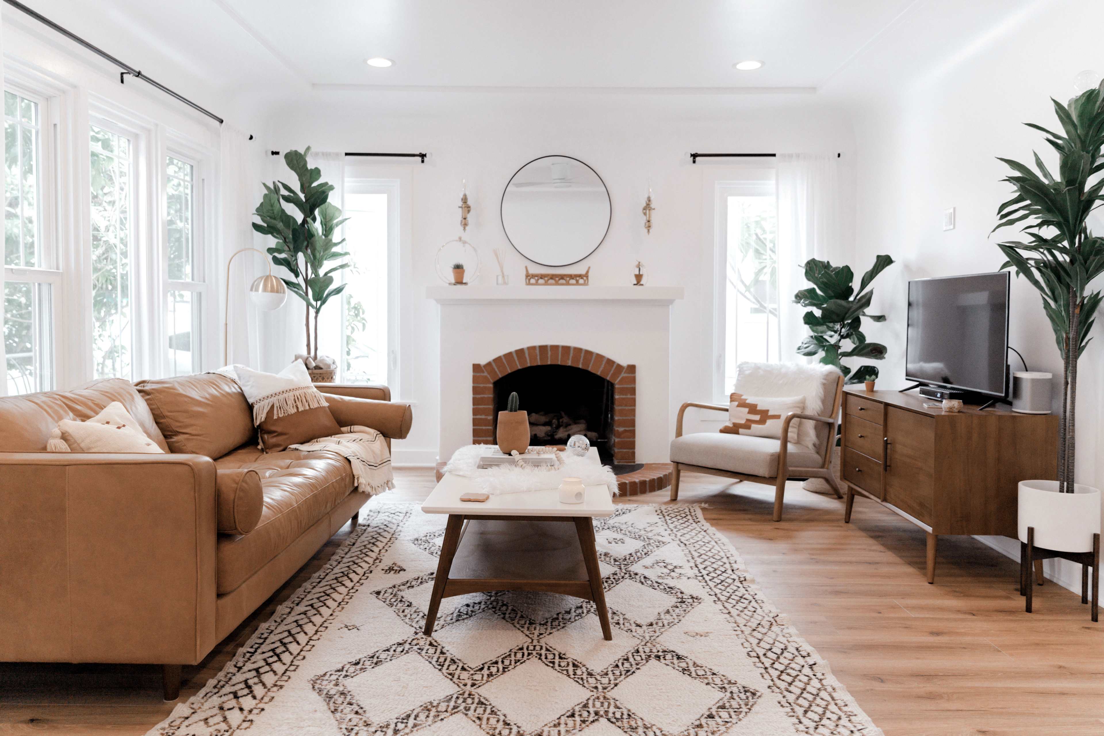 The living room features a tan leather sofa, a wooden coffee table on a patterned rug, and a fireplace surrounded by large windows, greenery, and a television.