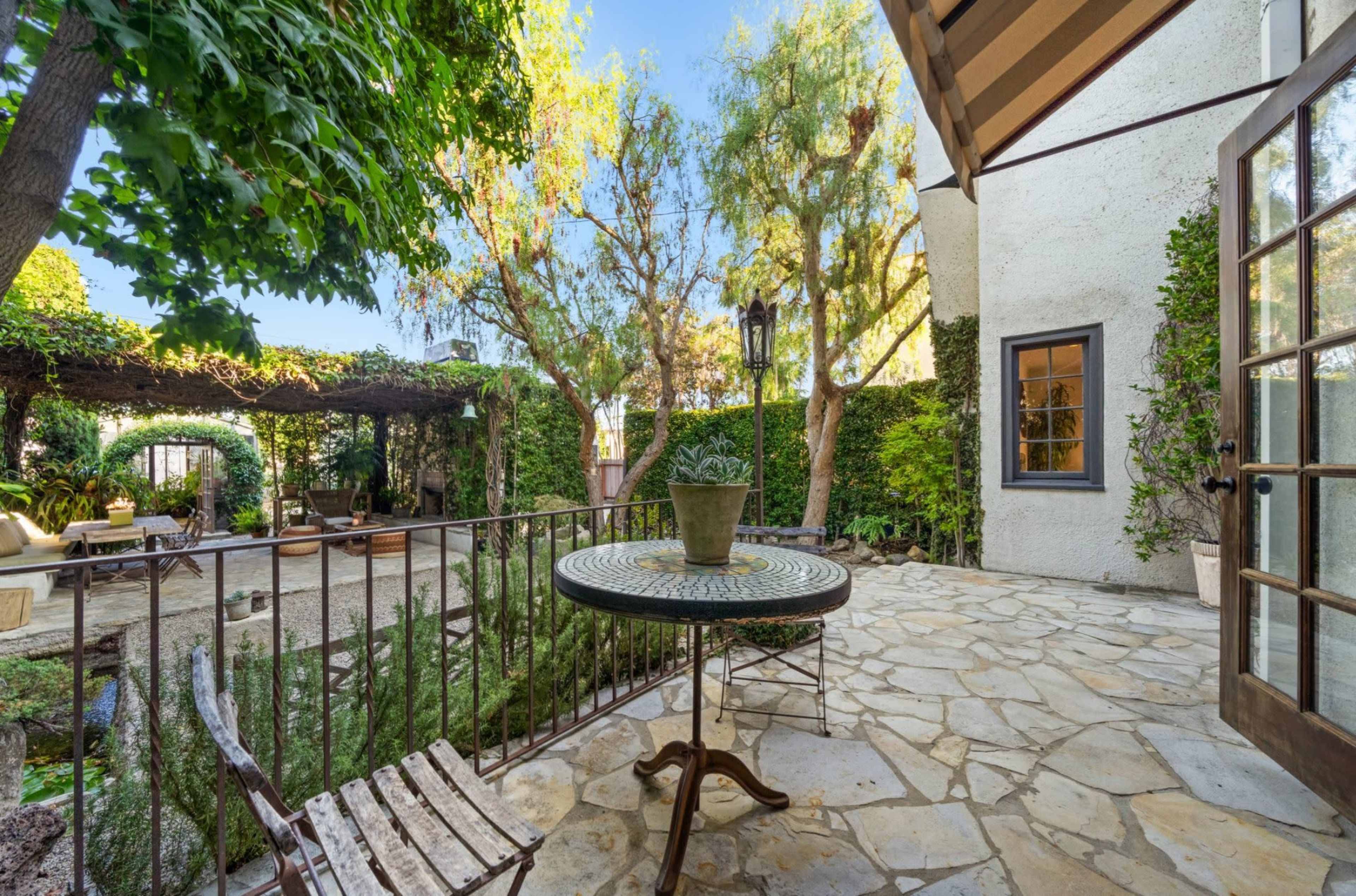 The image shows a stone patio with a small table and chair, surrounded by lush greenery and a view of a garden area.