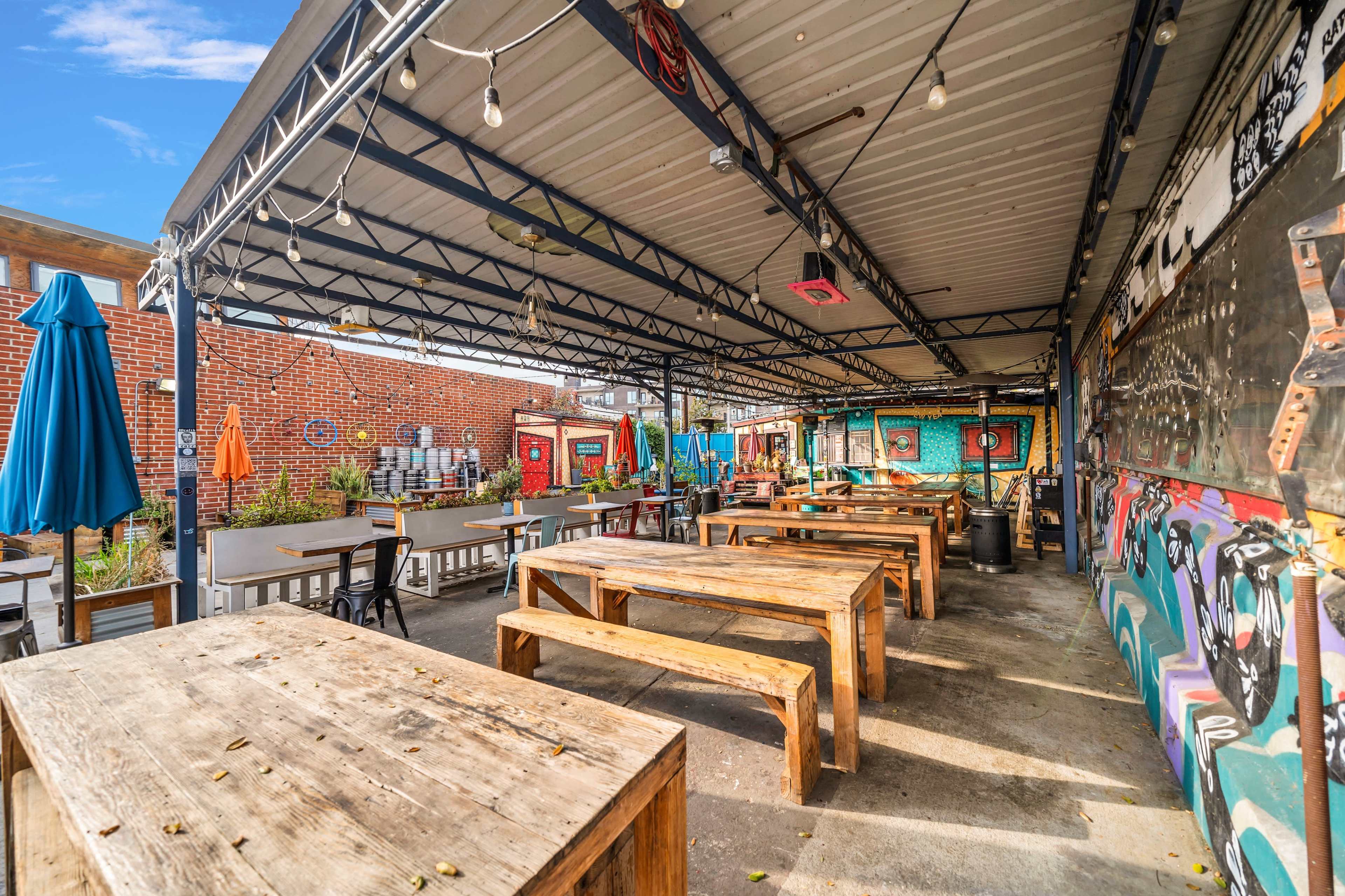 The image shows an outdoor dining area featuring wooden picnic tables beneath a covered structure, with colorful murals and string lights in the background.