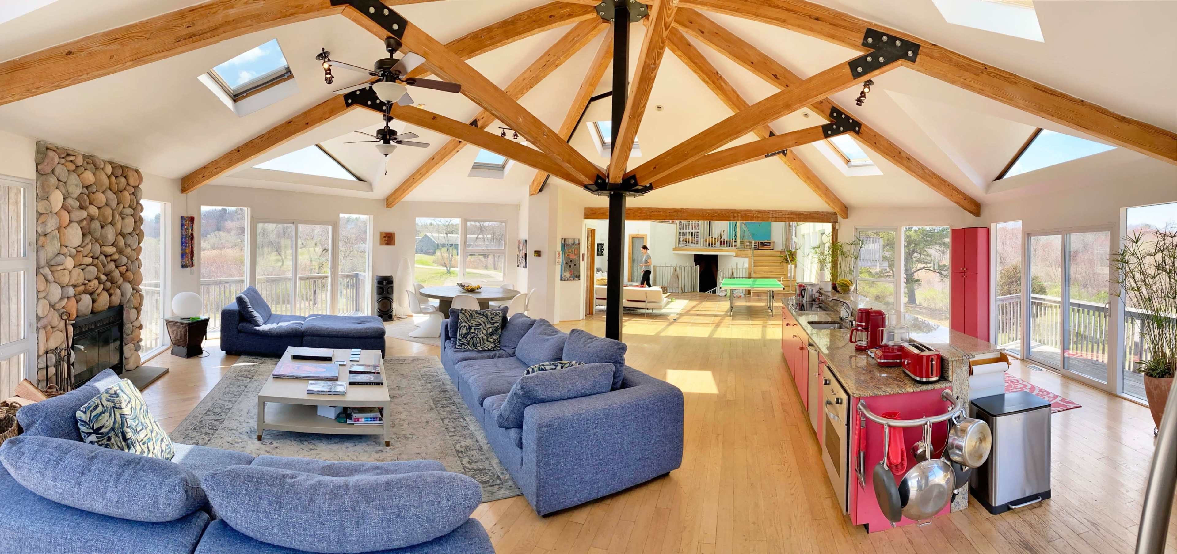 A spacious, modern living area with high wooden beams, two large sofas arranged around a coffee table, and an open kitchen area with red cabinets.
