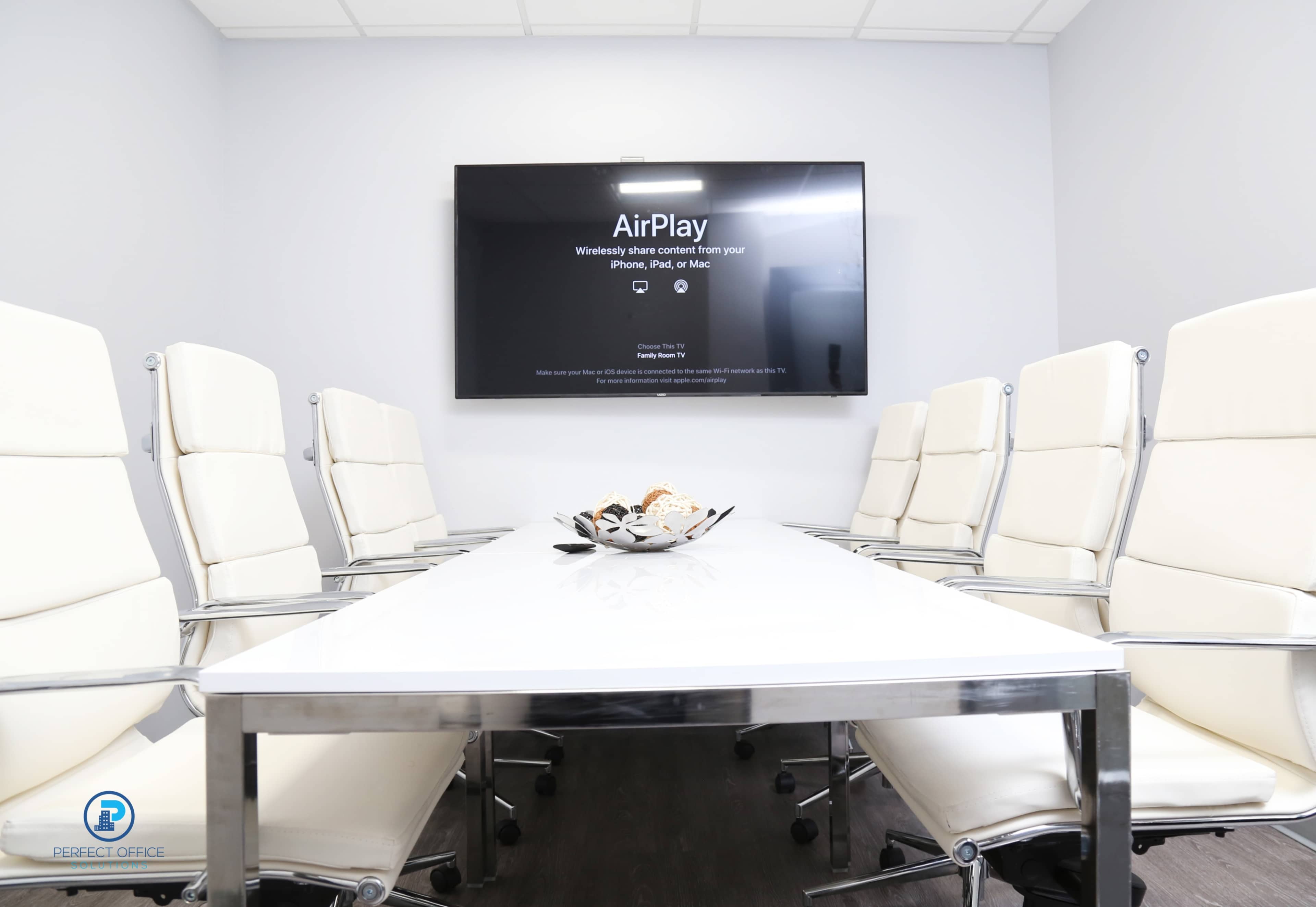 A modern conference room features a long white table surrounded by white chairs, with a wall-mounted screen displaying an AirPlay message.