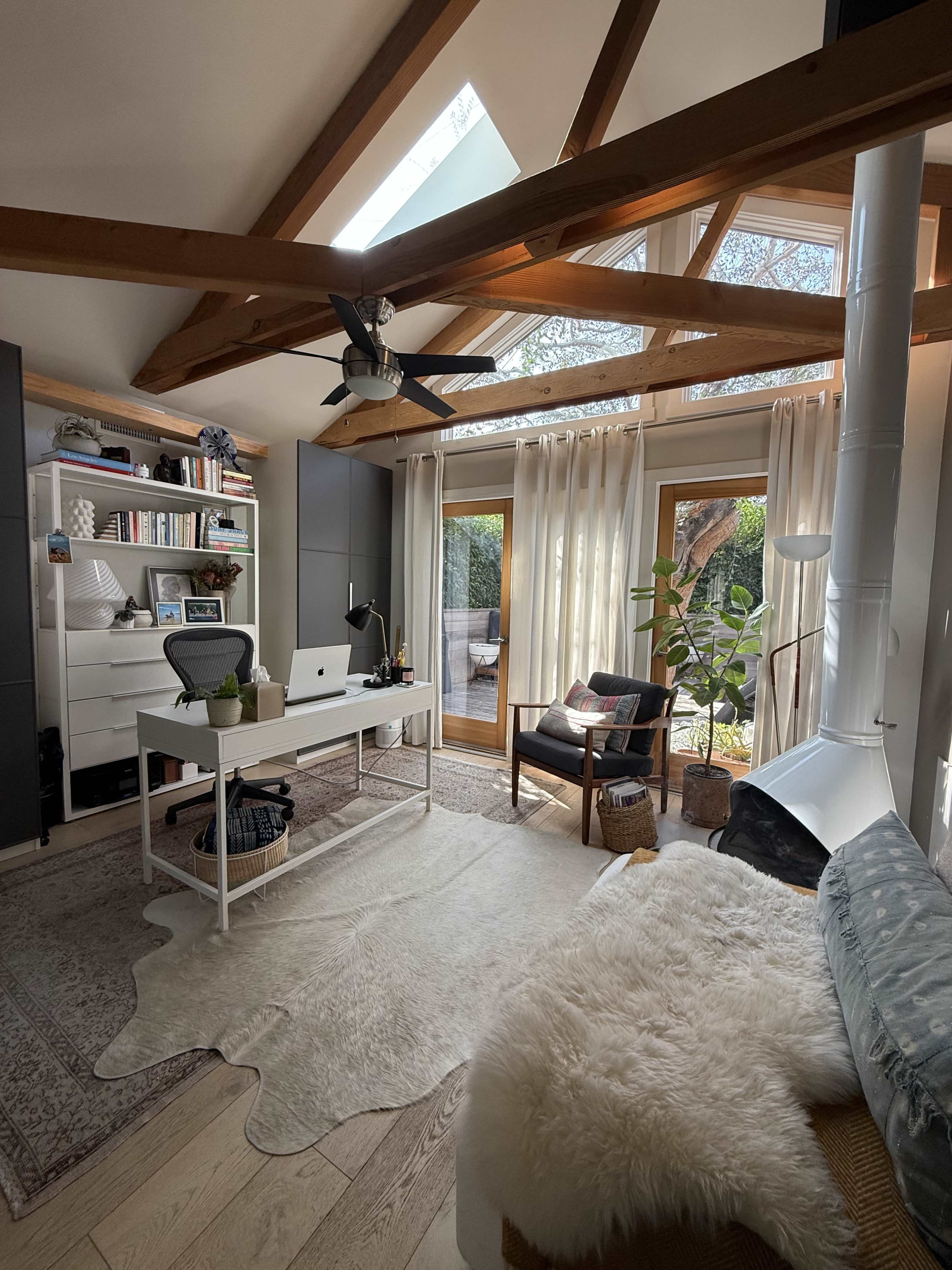 A stylish home office features a desk with a computer, surrounded by shelves and natural light pouring in through large windows beneath a wooden beam ceiling.