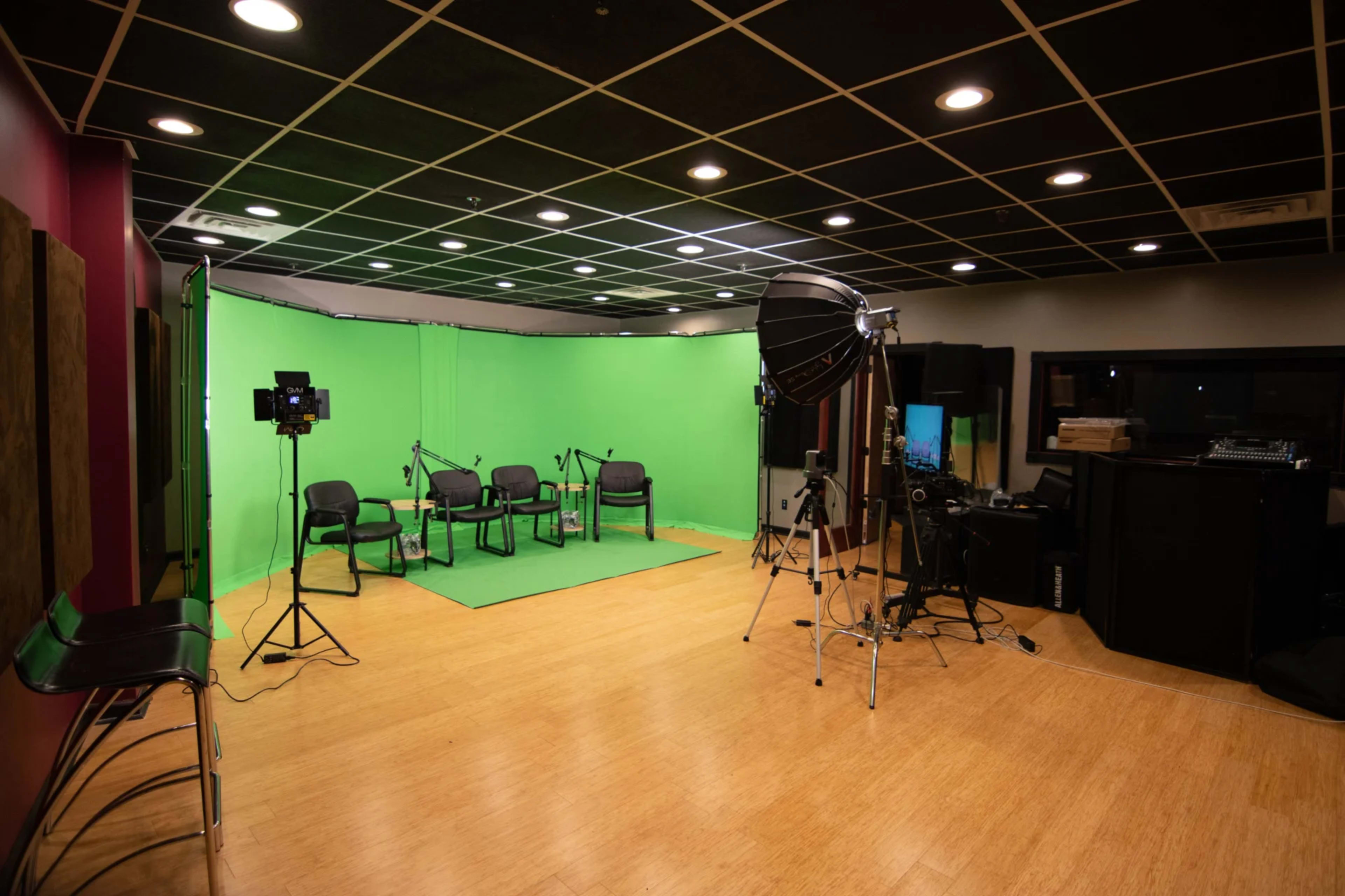The image shows a green screen studio setup with several chairs arranged, lights positioned, and camera equipment on the side.