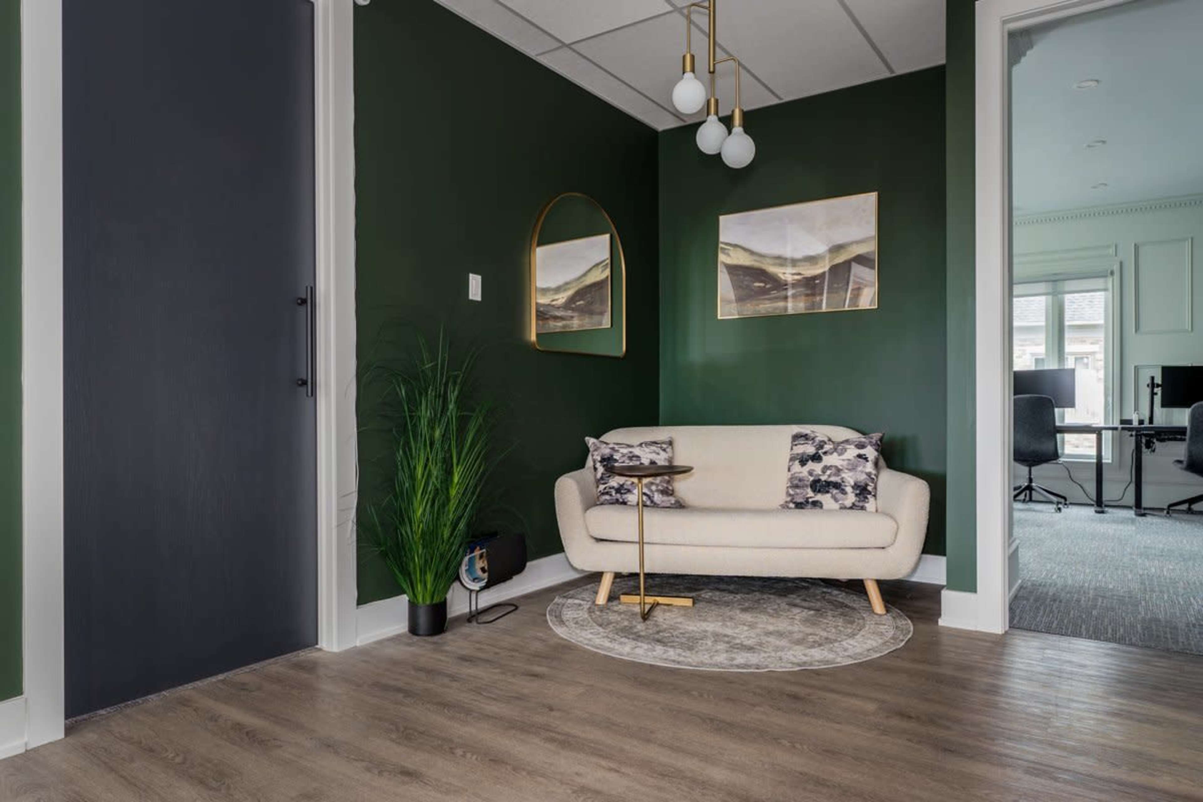 The image shows a cozy entryway featuring a green accent wall, a light-colored sofa with patterned cushions, and modern artwork, complemented by a plant and a round rug.