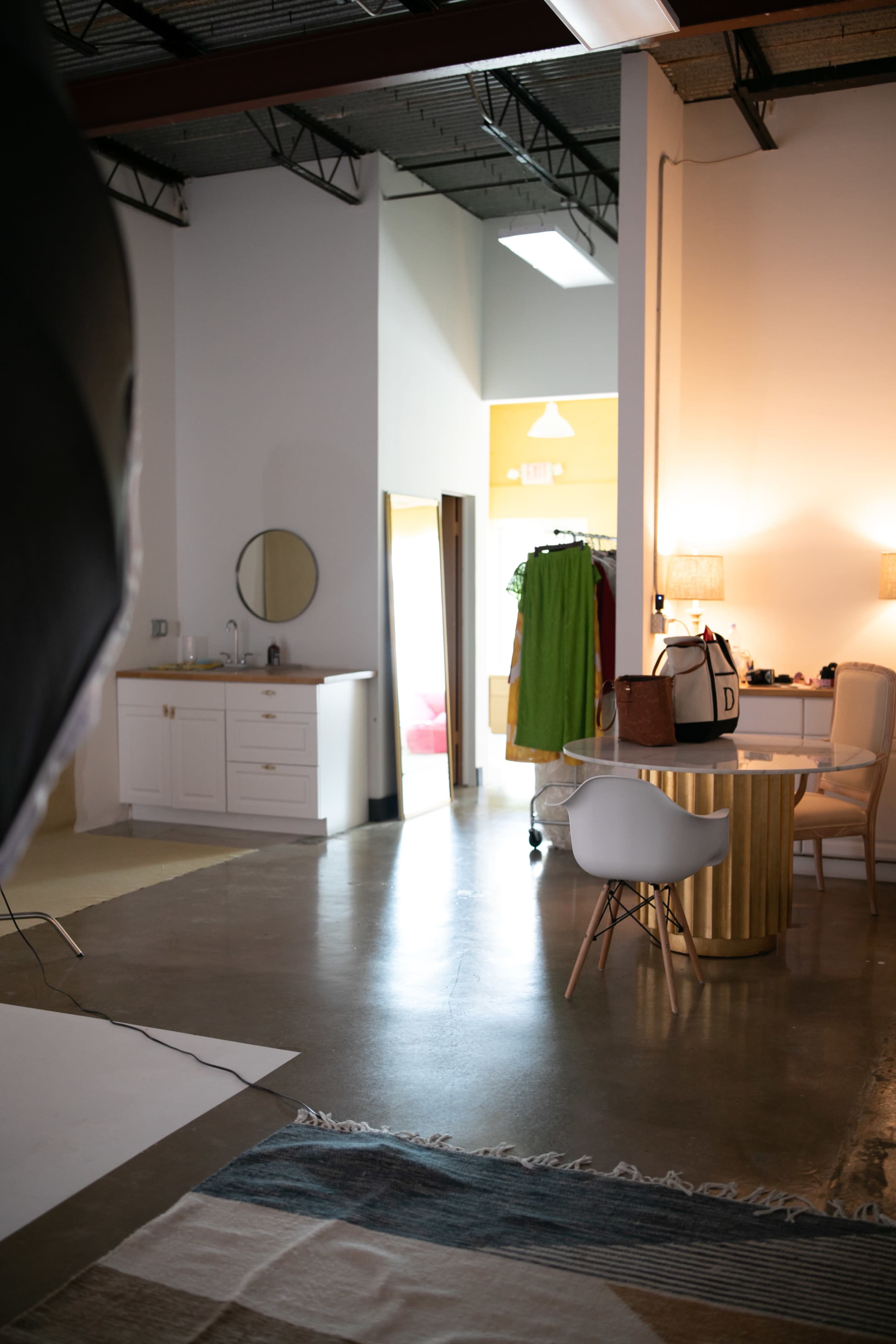 The image shows a studio space featuring a bathroom area, a clothing rack, a desk with a chair, and soft lighting.