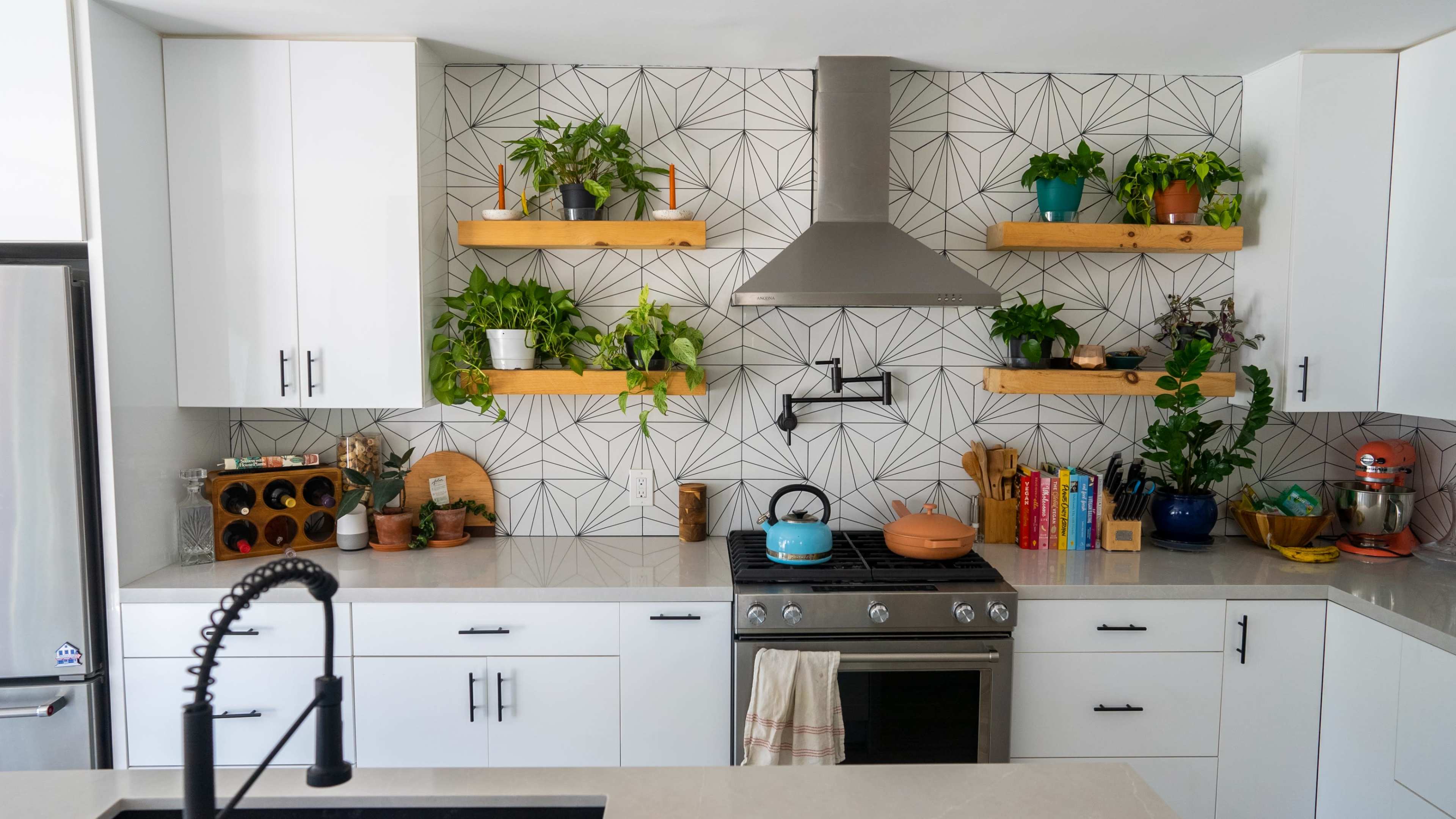 The kitchen features white cabinets, a stainless steel stove, and open shelves adorned with various potted plants and cookbooks against a geometric tile backdrop.