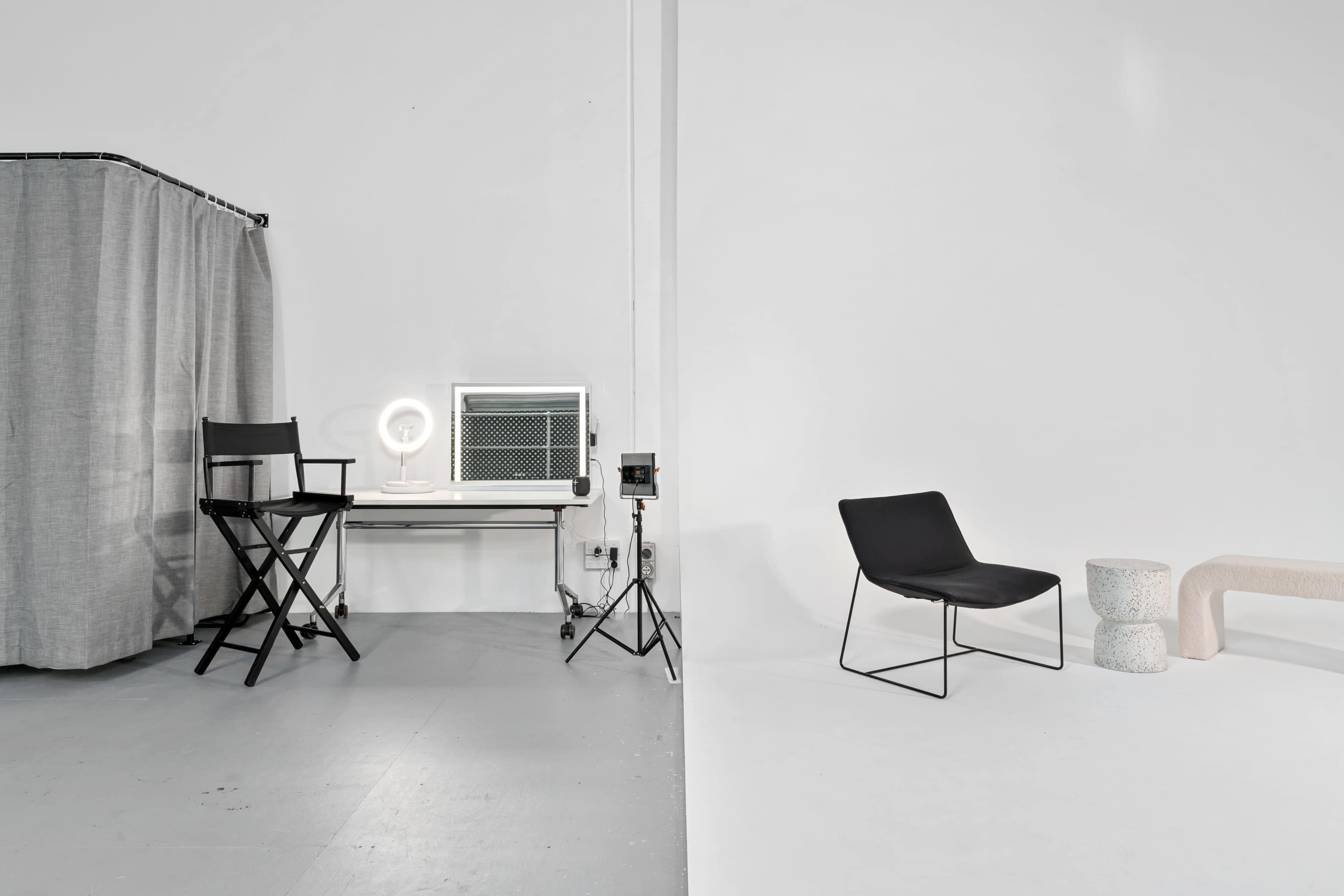 A minimalist studio space features a director's chair, a vanity with a ring light, and various seating options on opposite sides of a divided area.