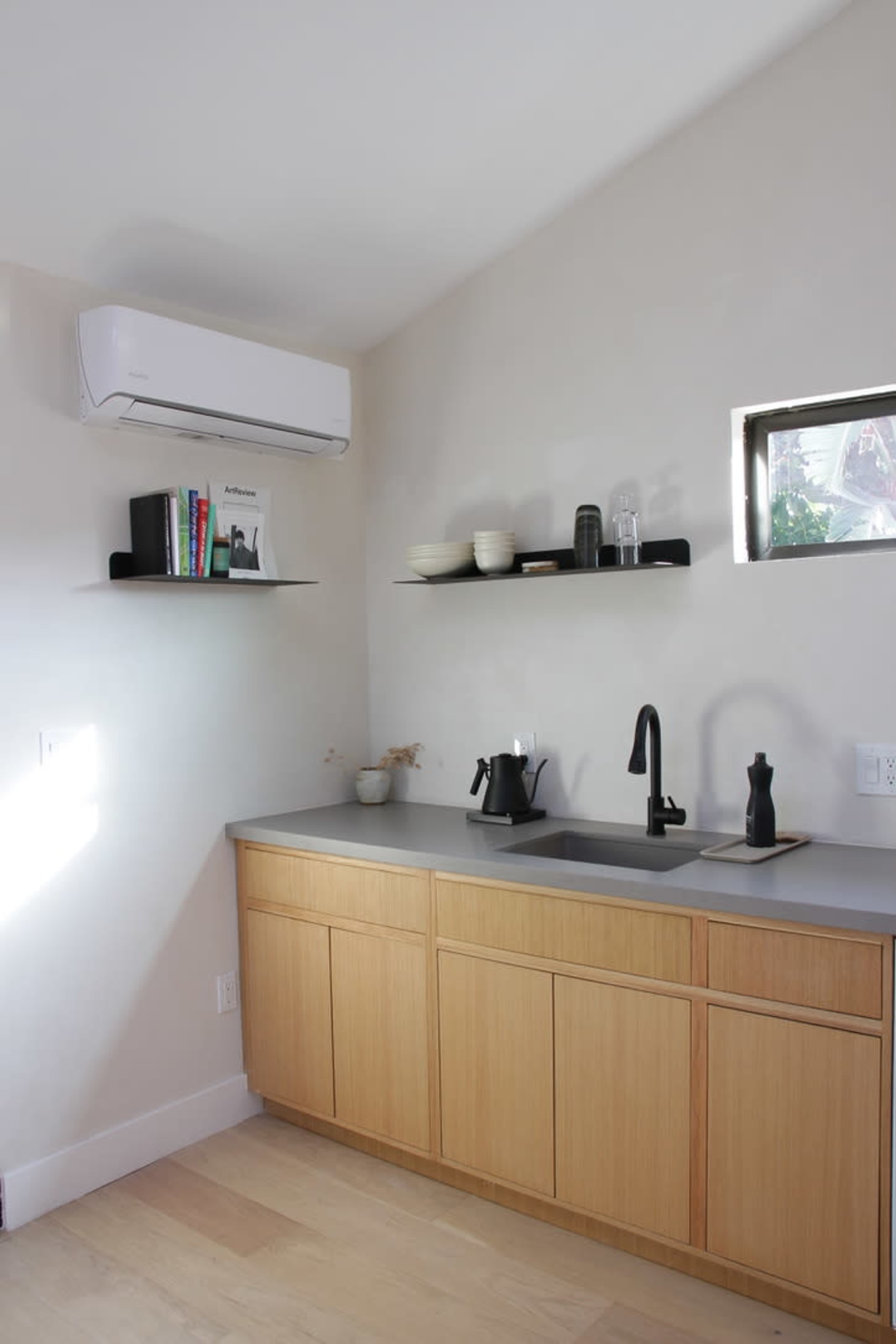 A modern kitchen with a wooden cabinet under a gray countertop, featuring a black faucet and a small shelf displaying books and dishware.