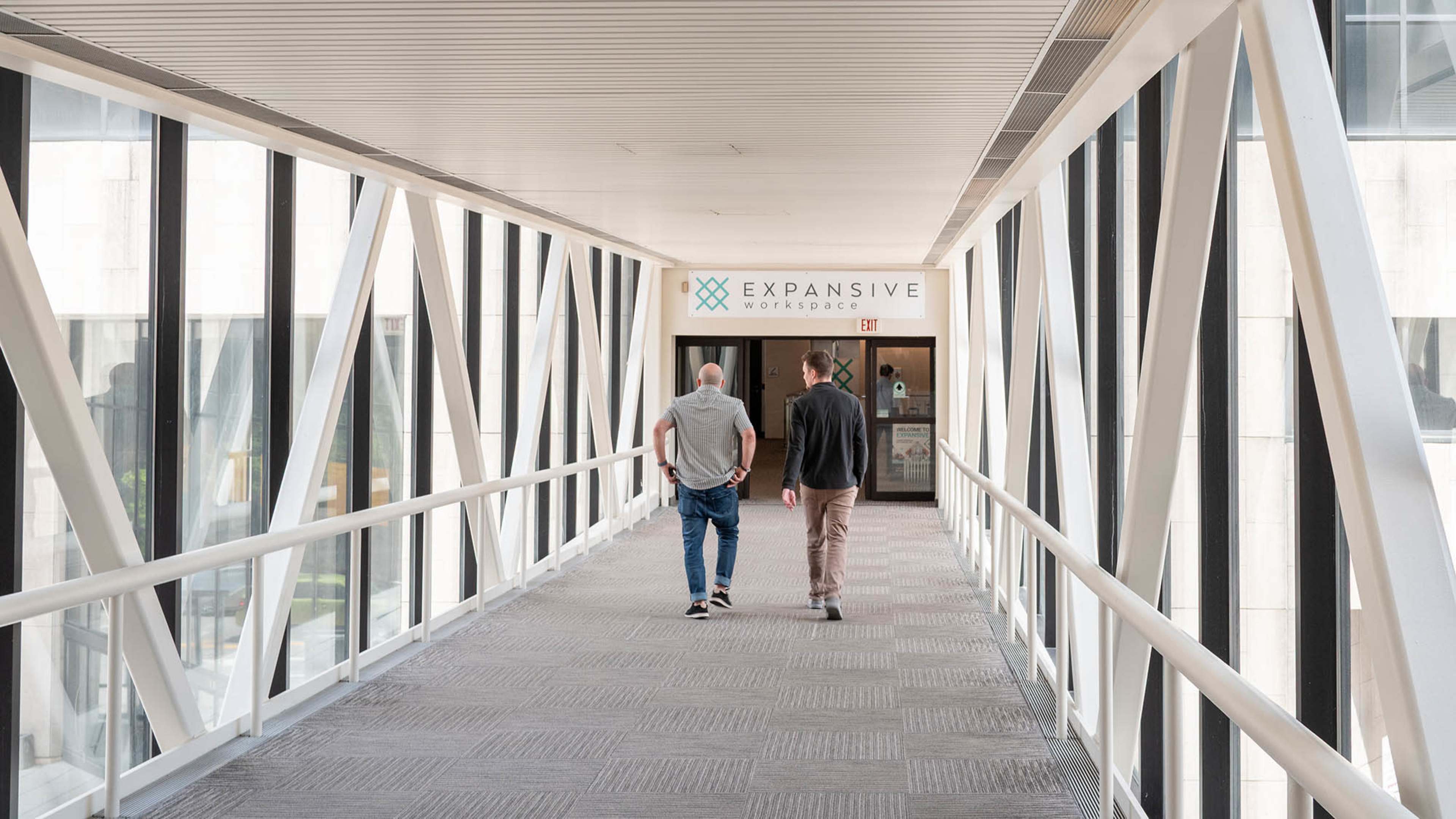 Two men walk down a modern corridor with a sign reading "Expansive Workspace" at the entrance.