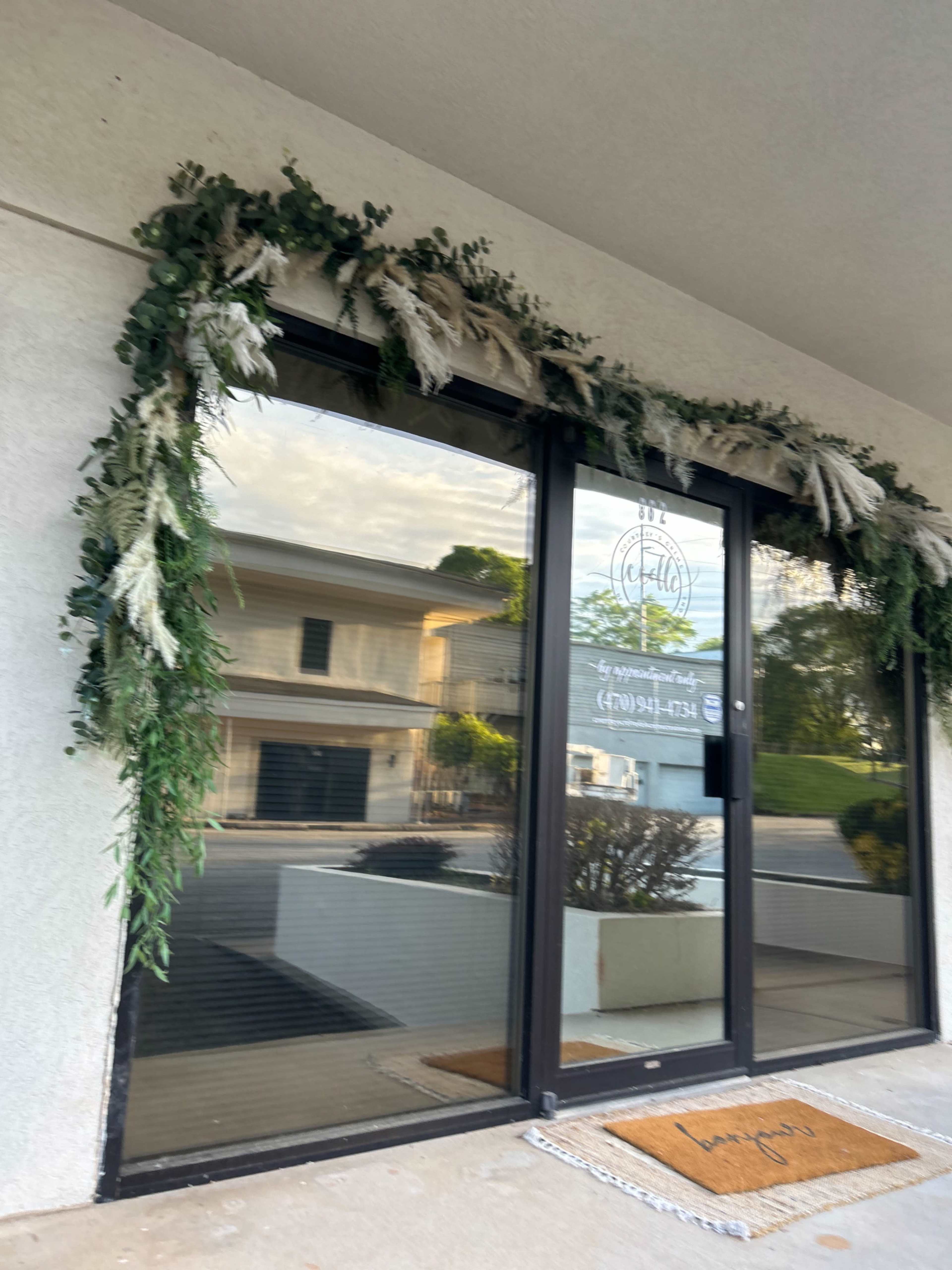 The exterior of a storefront features large glass doors adorned with a greenery and dried flower garland.