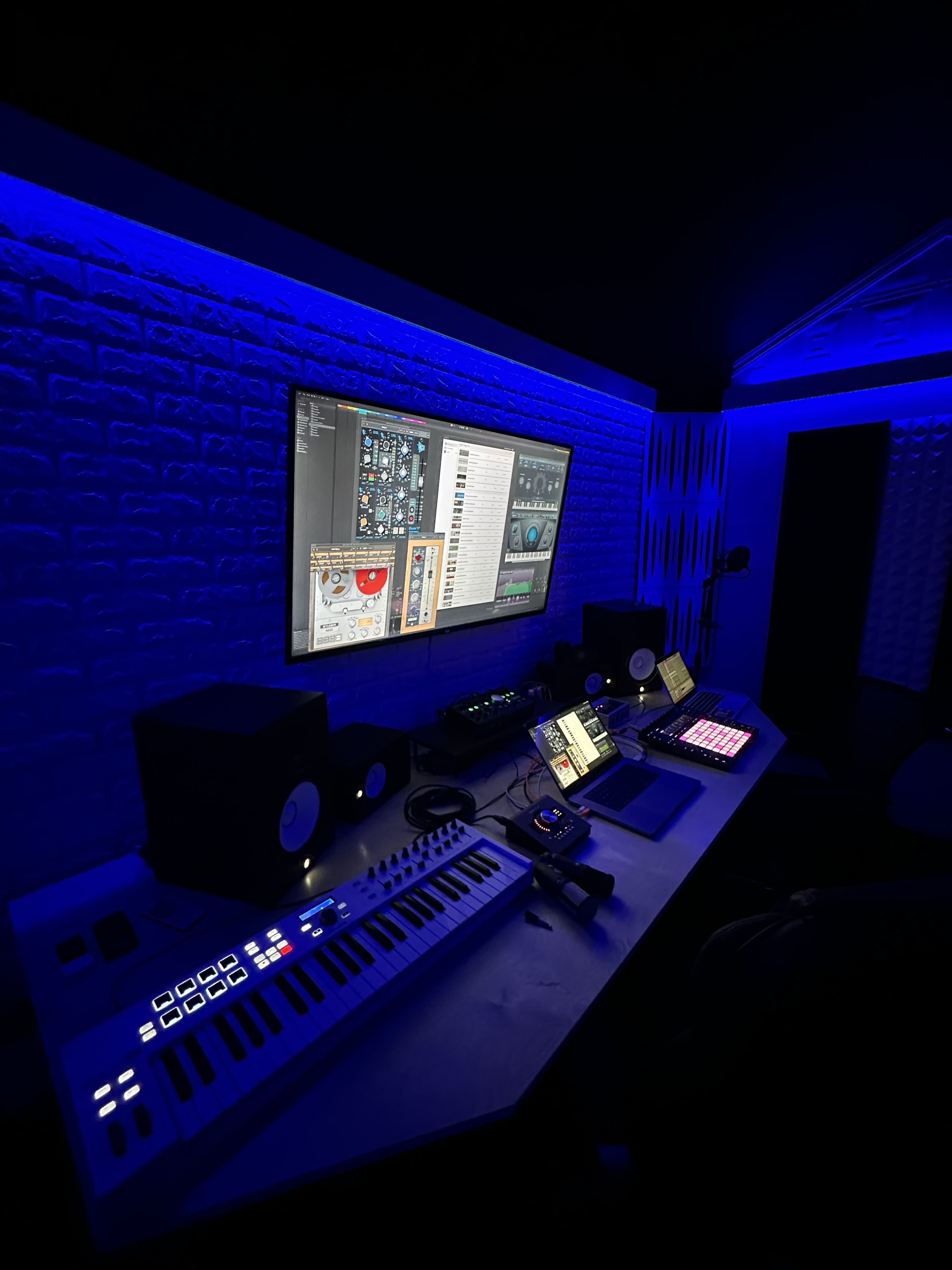 A music production studio is illuminated with blue lighting, featuring a computer monitor displaying audio software, keyboards, and various music equipment on the desk.