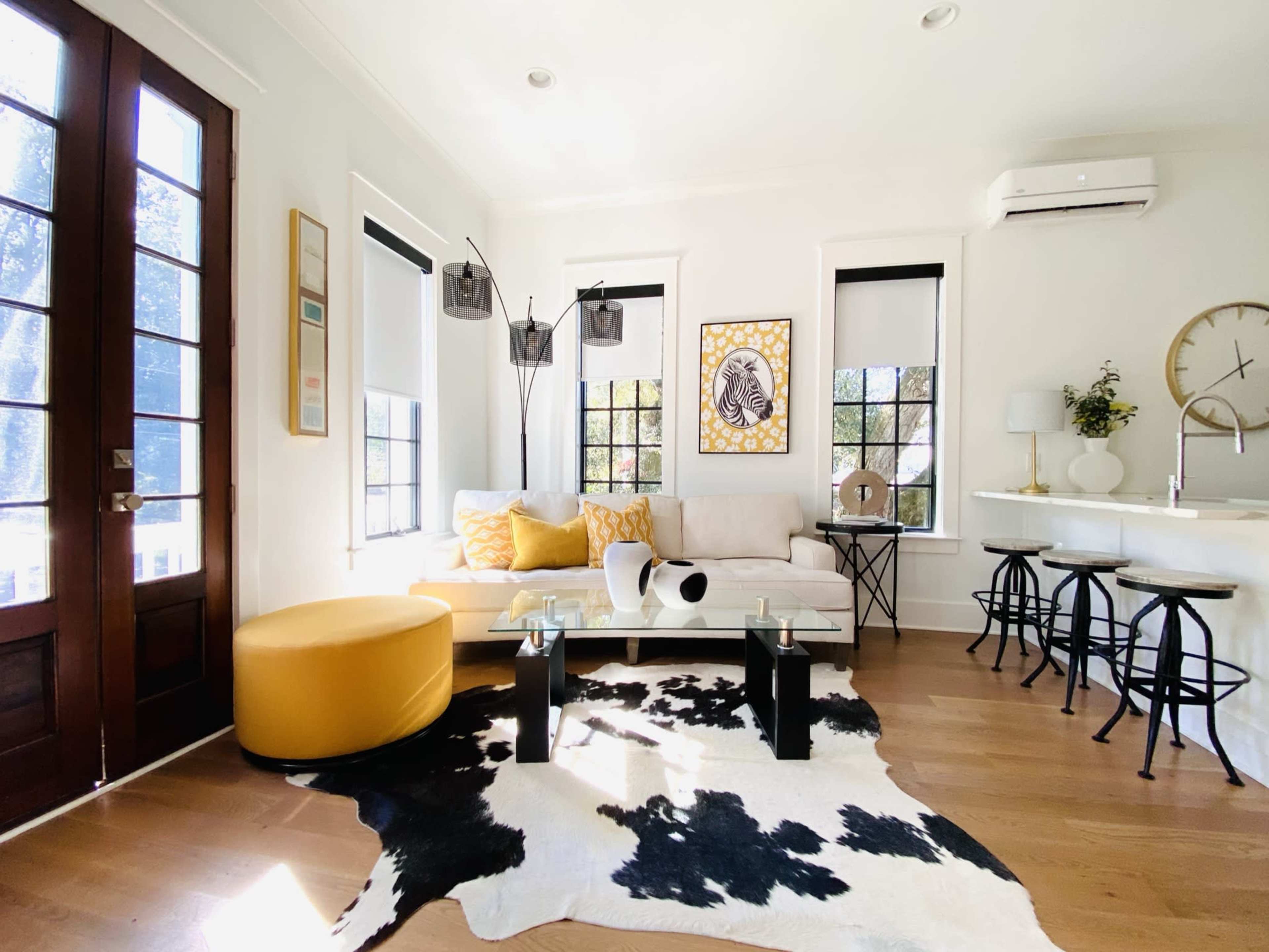 A modern living room features a white sofa with yellow accents, a glass coffee table on a cowhide rug, and large windows letting in natural light.