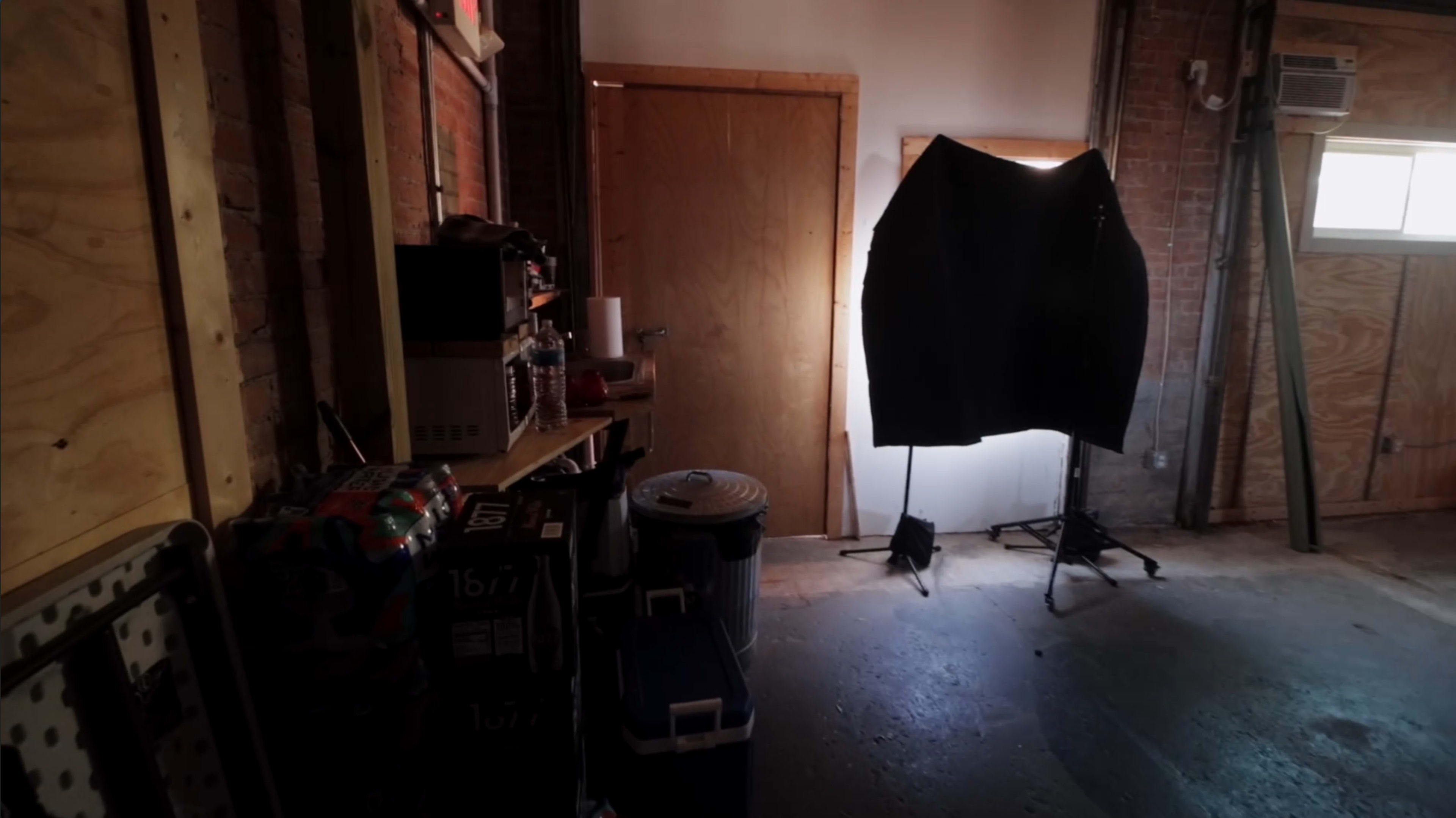 A dimly lit room with wooden walls, a closed door, a black backdrop on a stand, and various equipment and containers lining a shelf.