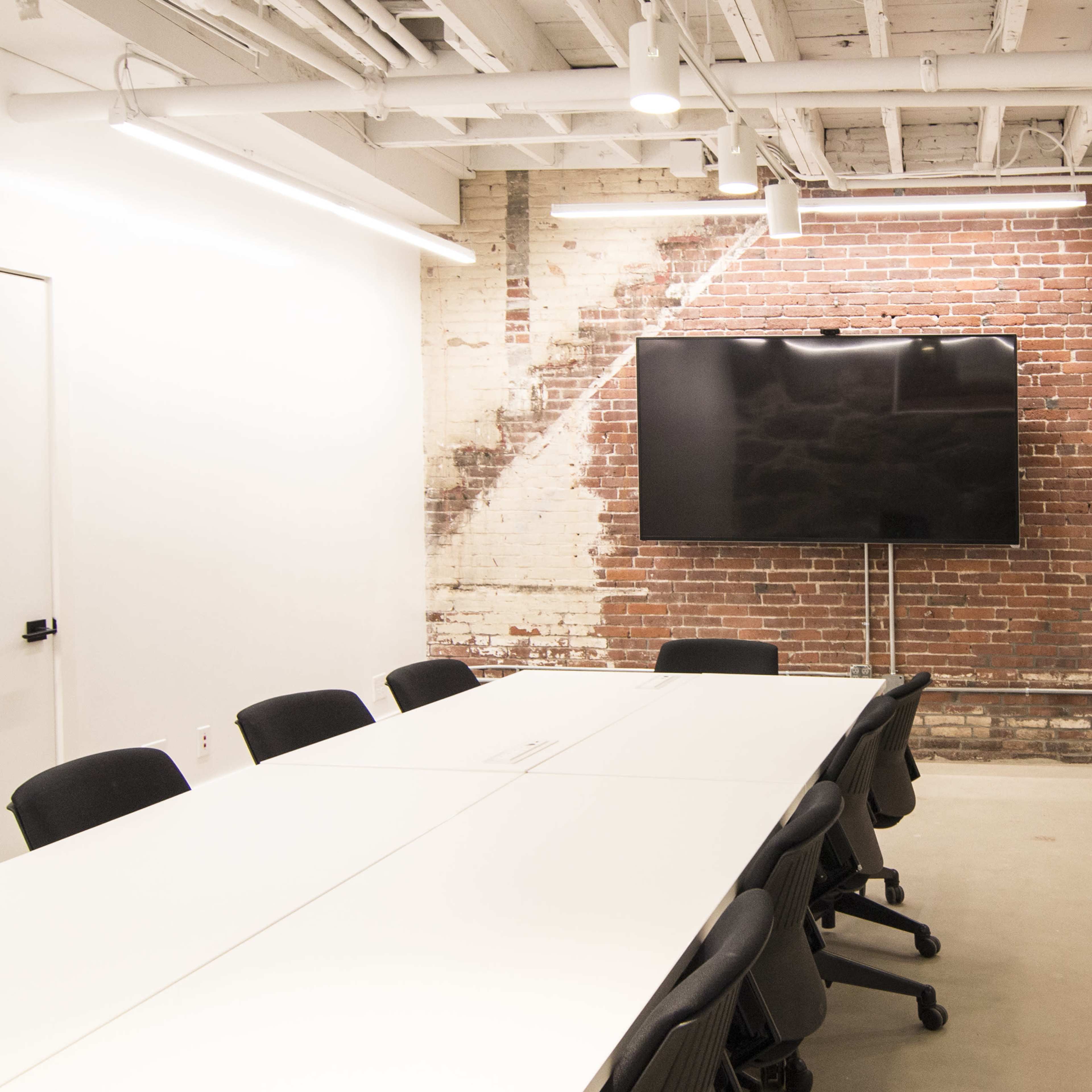 A modern conference room features a long white table surrounded by black chairs and a large television mounted on a brick wall.