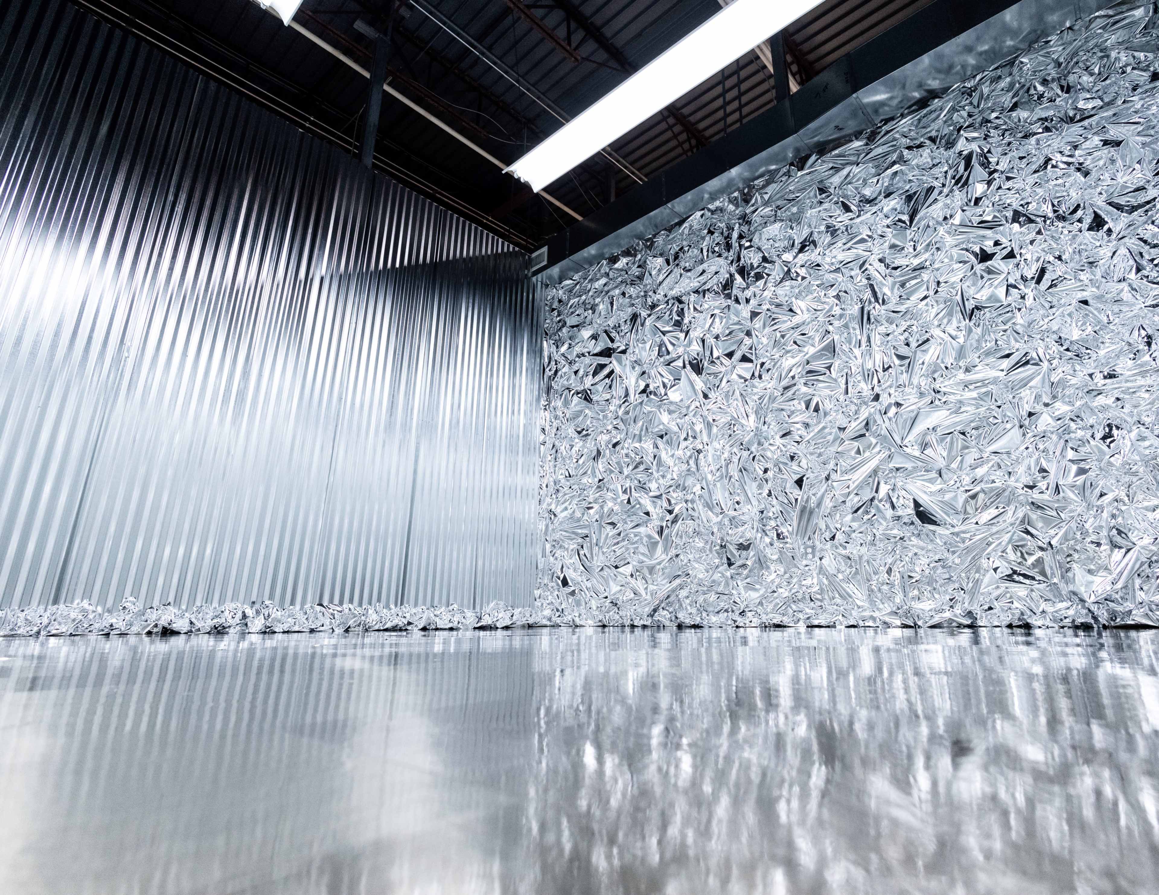 A reflective metallic surface with a wall made of crumpled foil-like material and a backdrop of corrugated metal panels.