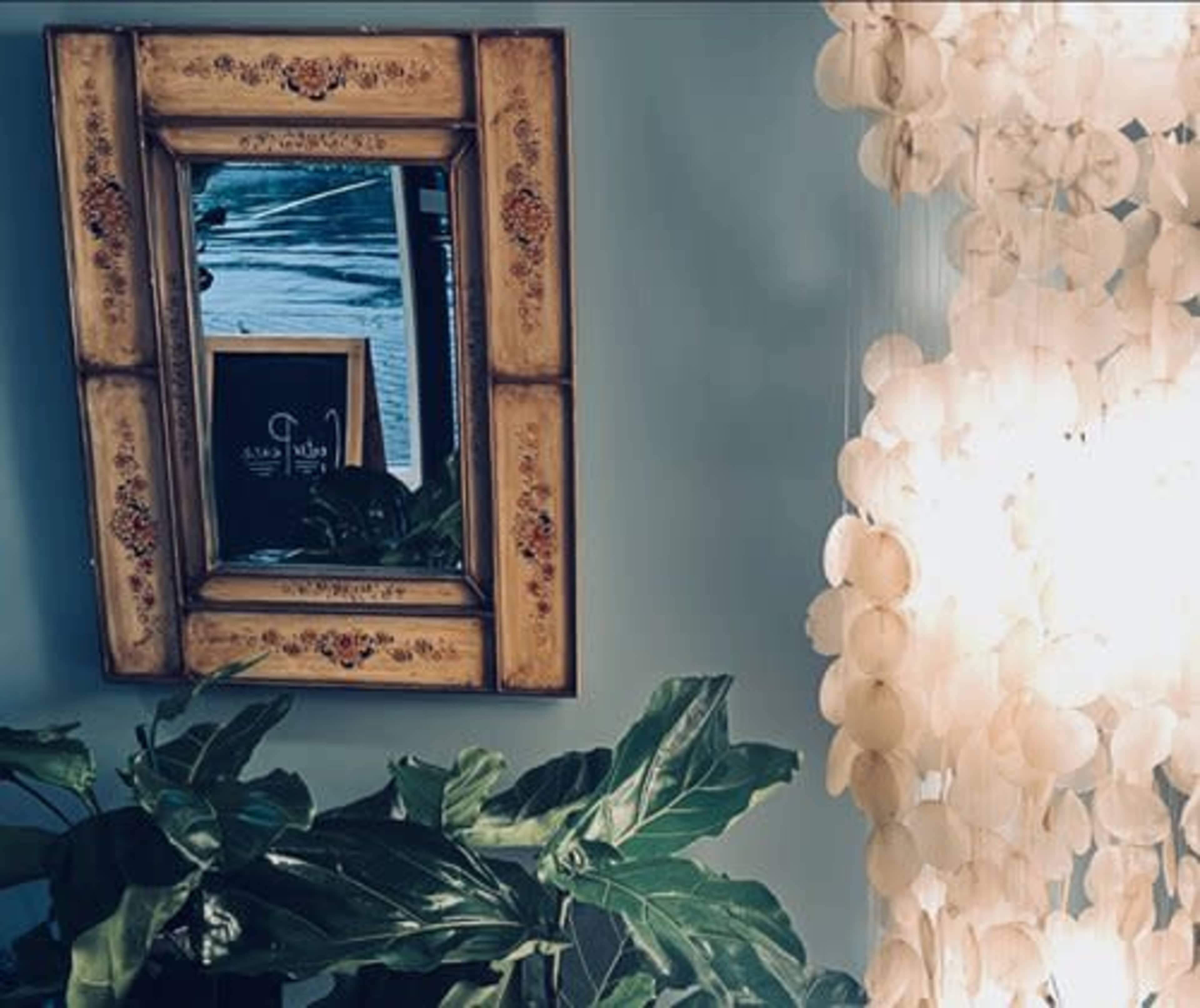 A decorative mirror with a floral frame reflects a softly lit room and is surrounded by lush green plants and a curtain of circular decorative elements.