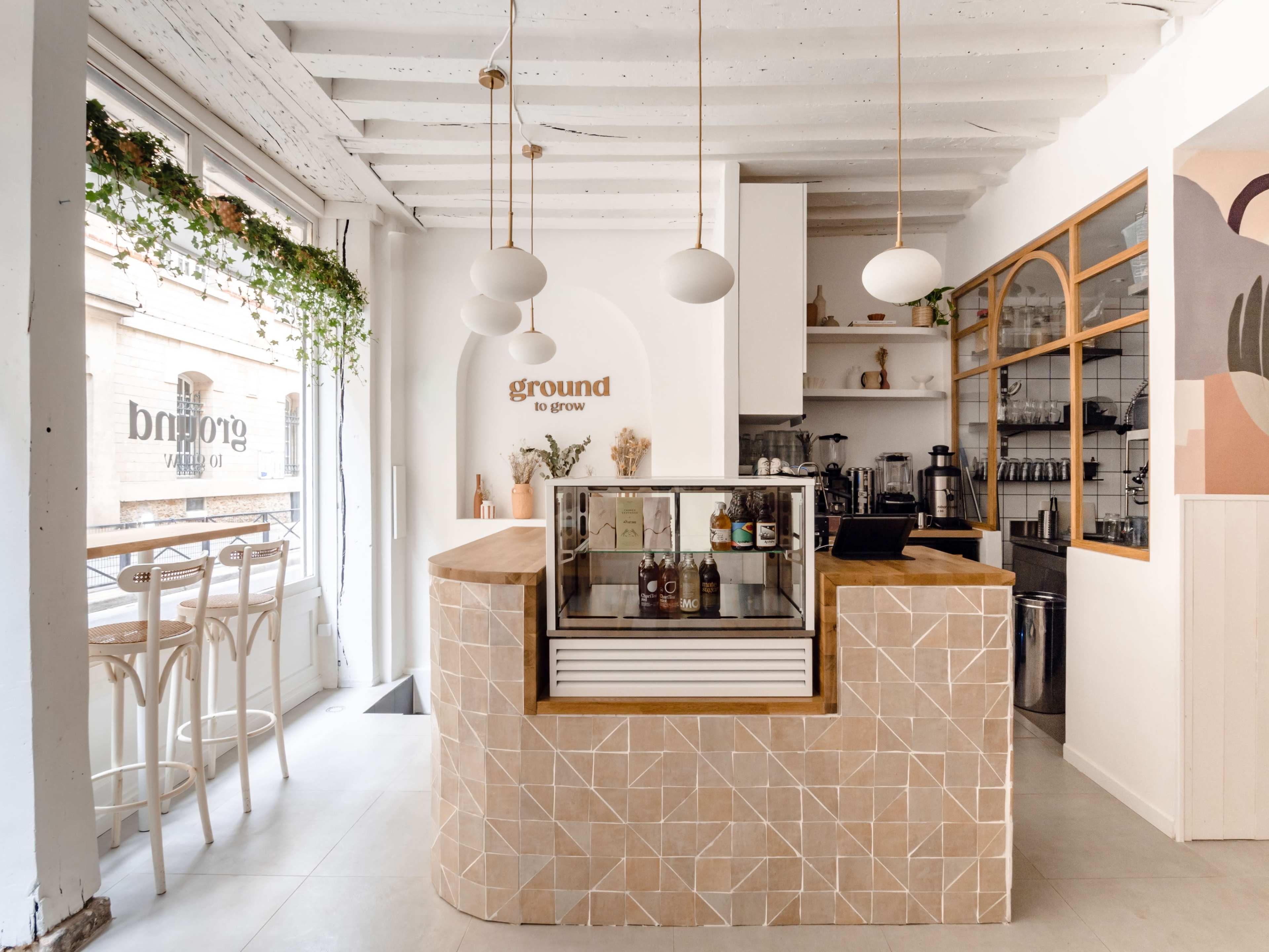 The image shows a modern café interior featuring a light-colored counter with a glass display, minimalistic decor, and barstool seating near large windows.