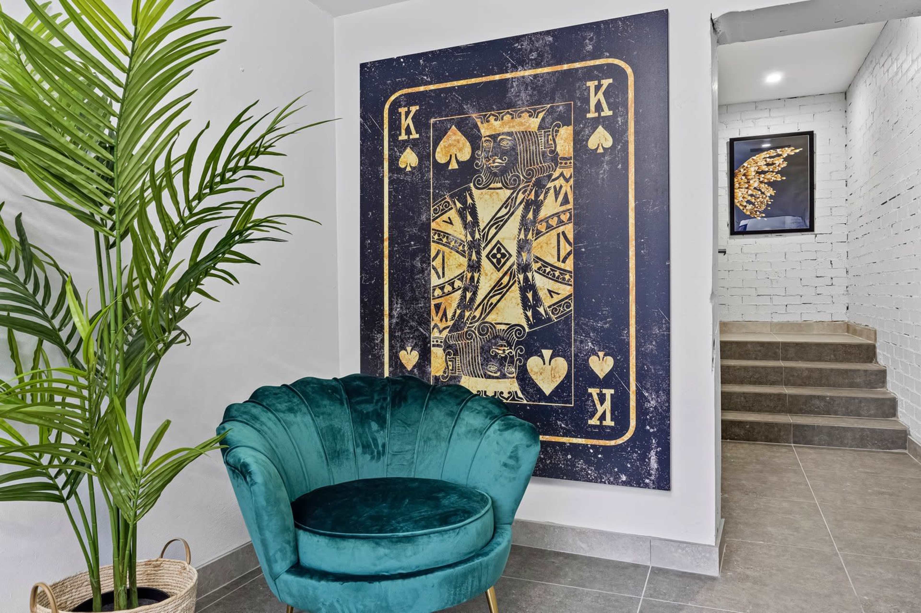 A large wall art piece featuring a King of Spades card hangs above a stylish teal velvet chair, with a potted plant nearby and stairs leading to another level.