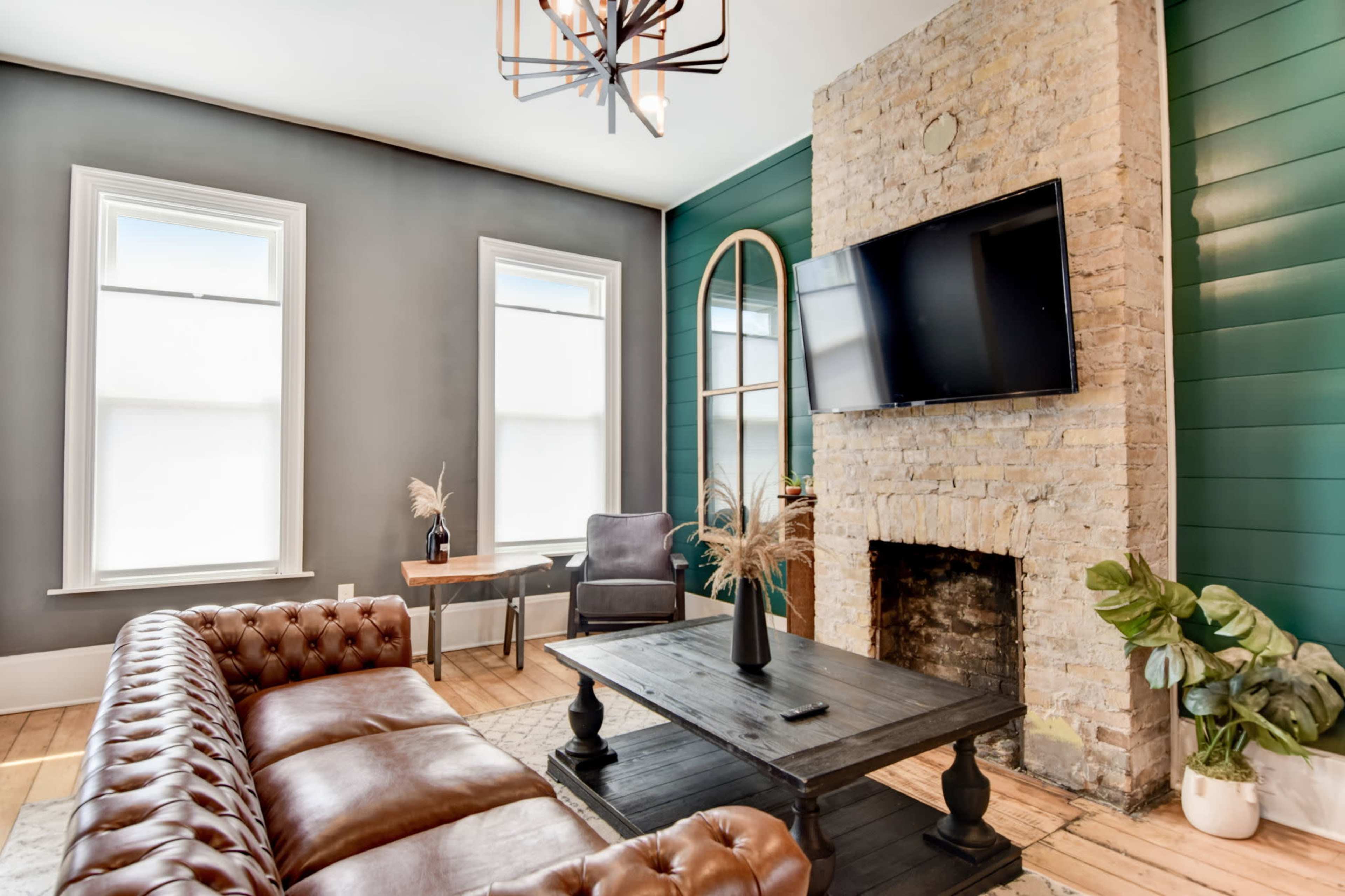 The room features a leather sofa, a stone fireplace, a mounted television, and green accent walls with large windows.
