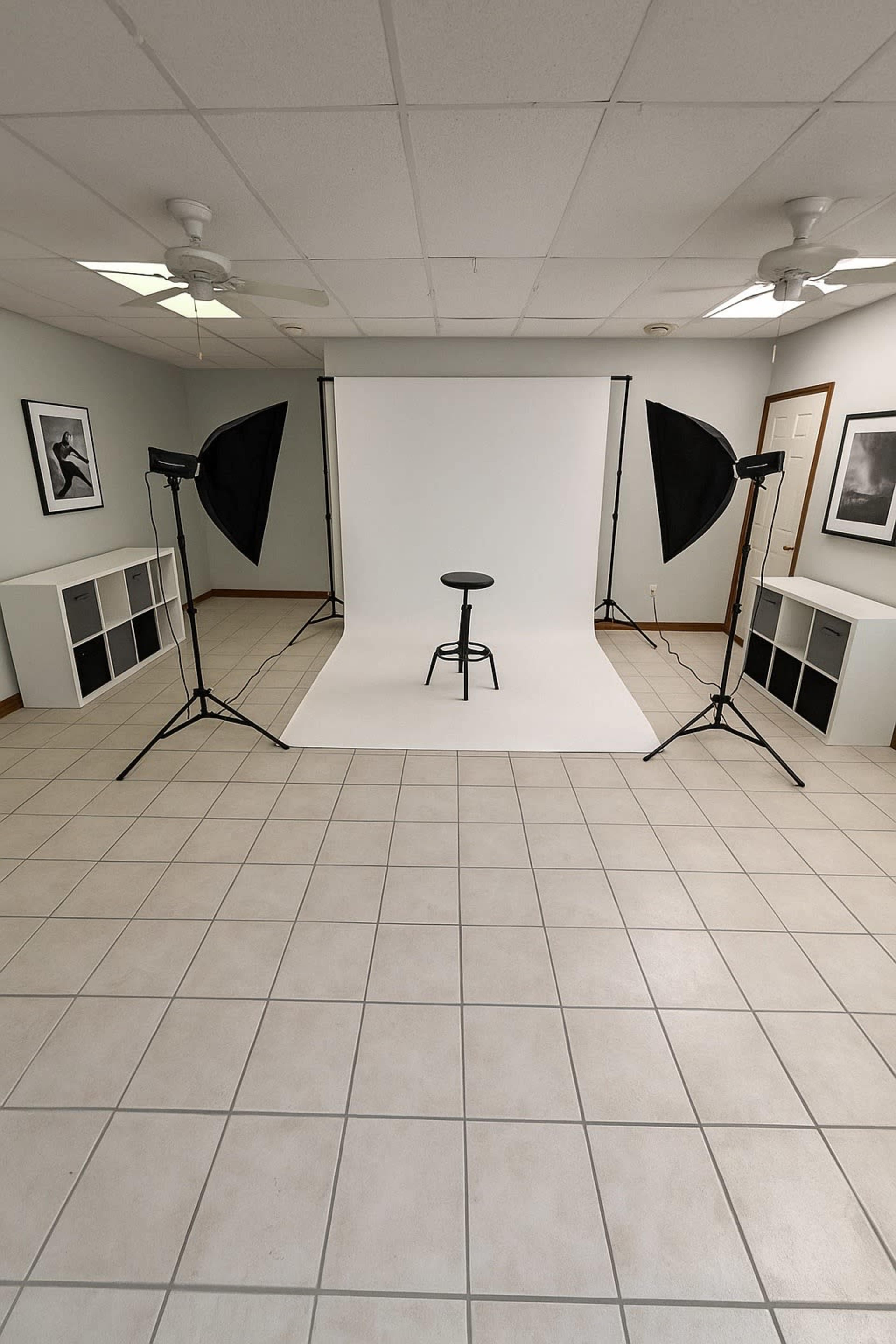 The image shows a photography studio setup with a white backdrop, a black stool in the center, and two softbox lights positioned on either side.