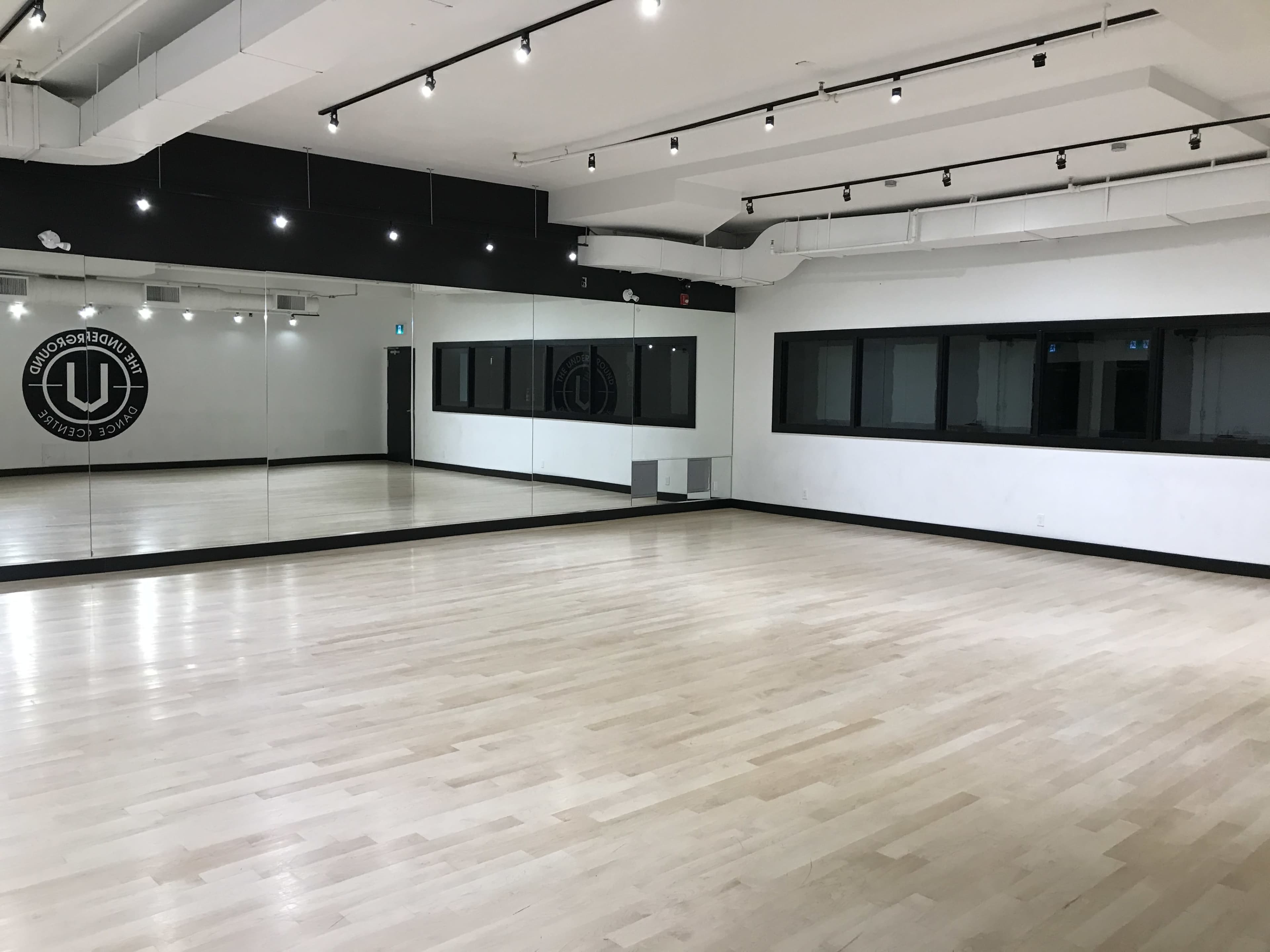 The image shows an empty dance studio with wooden flooring, mirrors on one wall, and large windows on another wall.