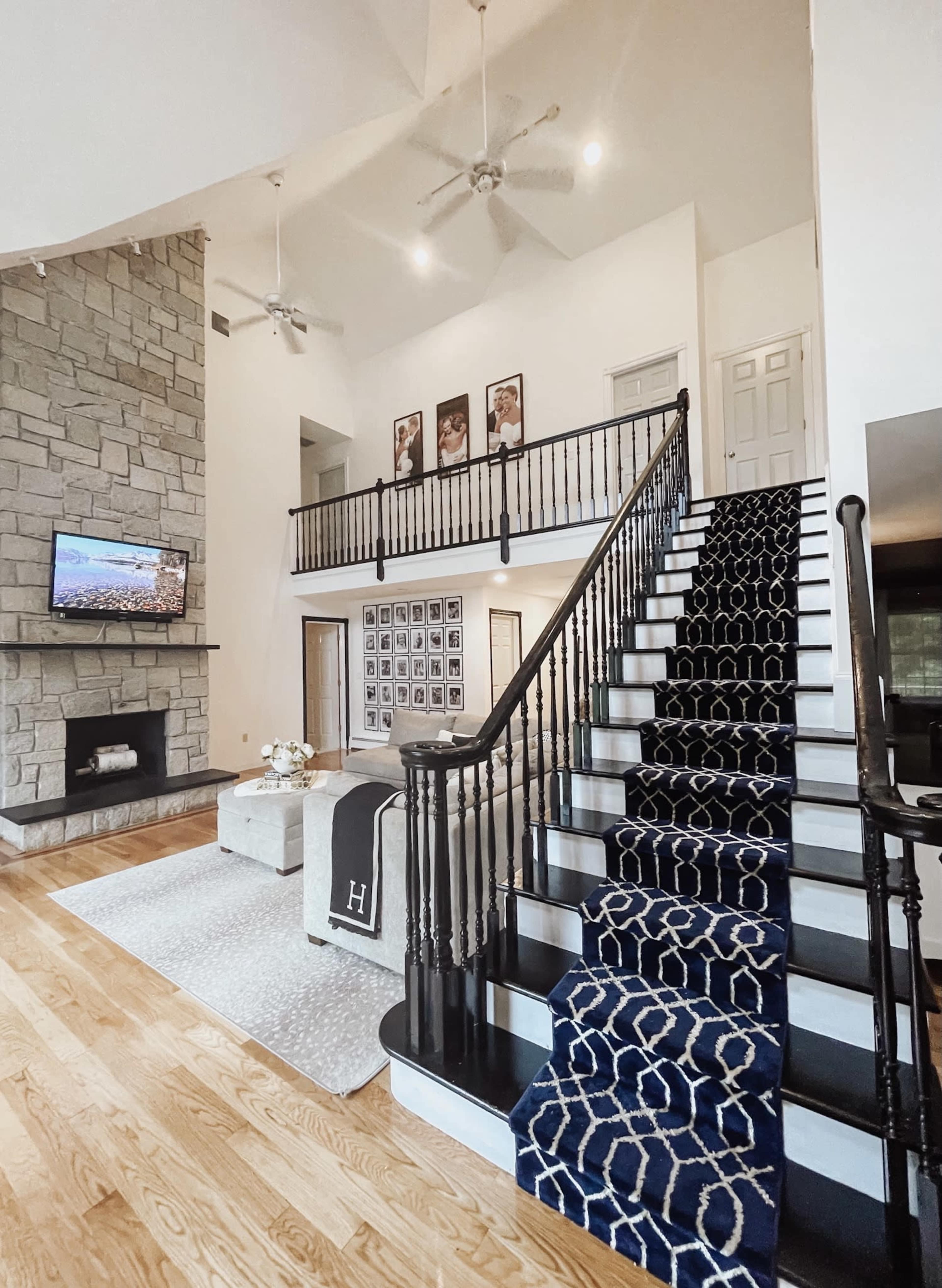 A spacious living area with a staircase featuring a patterned runner, a stone fireplace, and a wall adorned with black-and-white photographs.