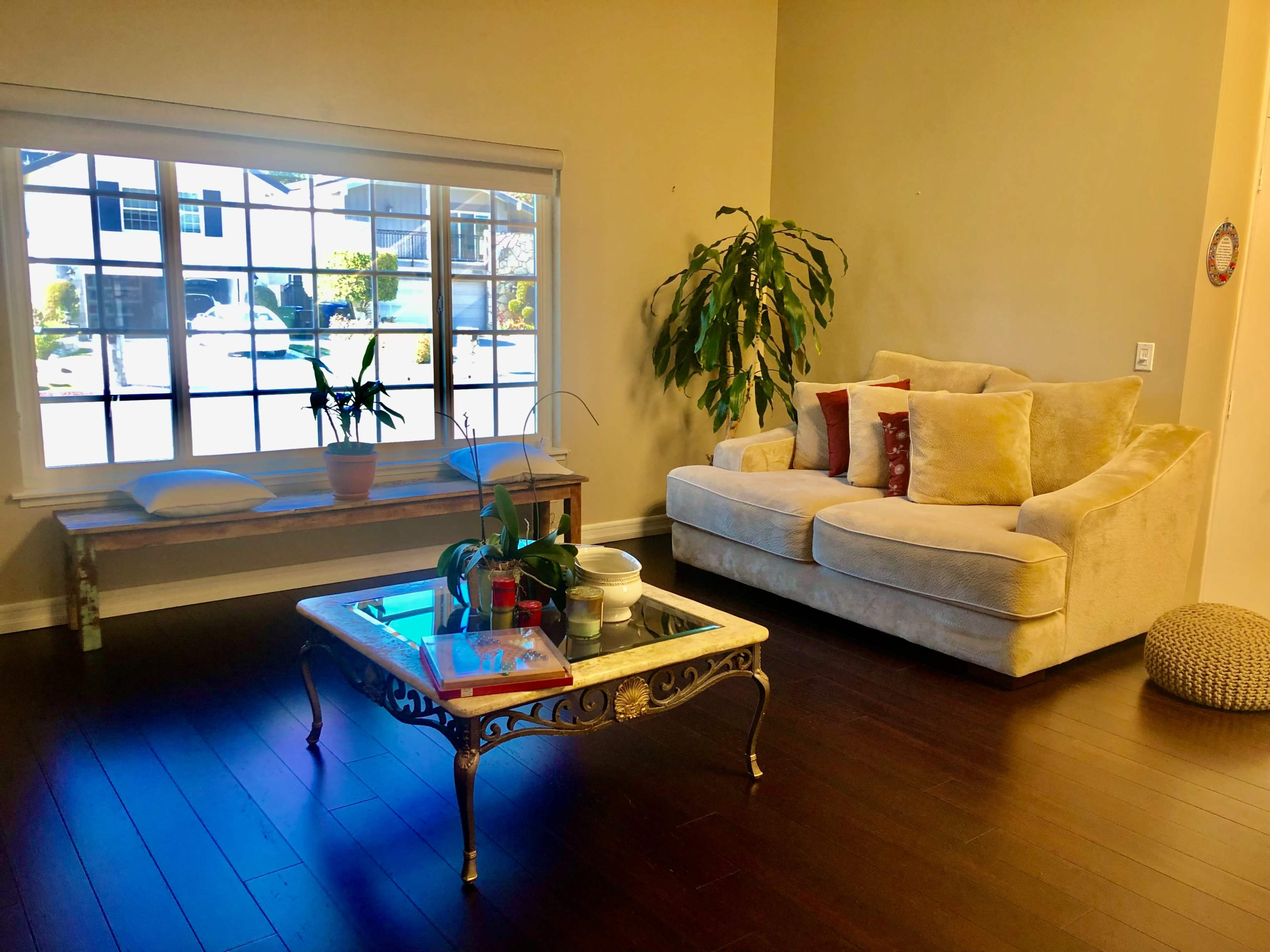 A cozy living room features a beige sofa, a coffee table with glass top, and a bench beneath a large window overlooking a residential street.