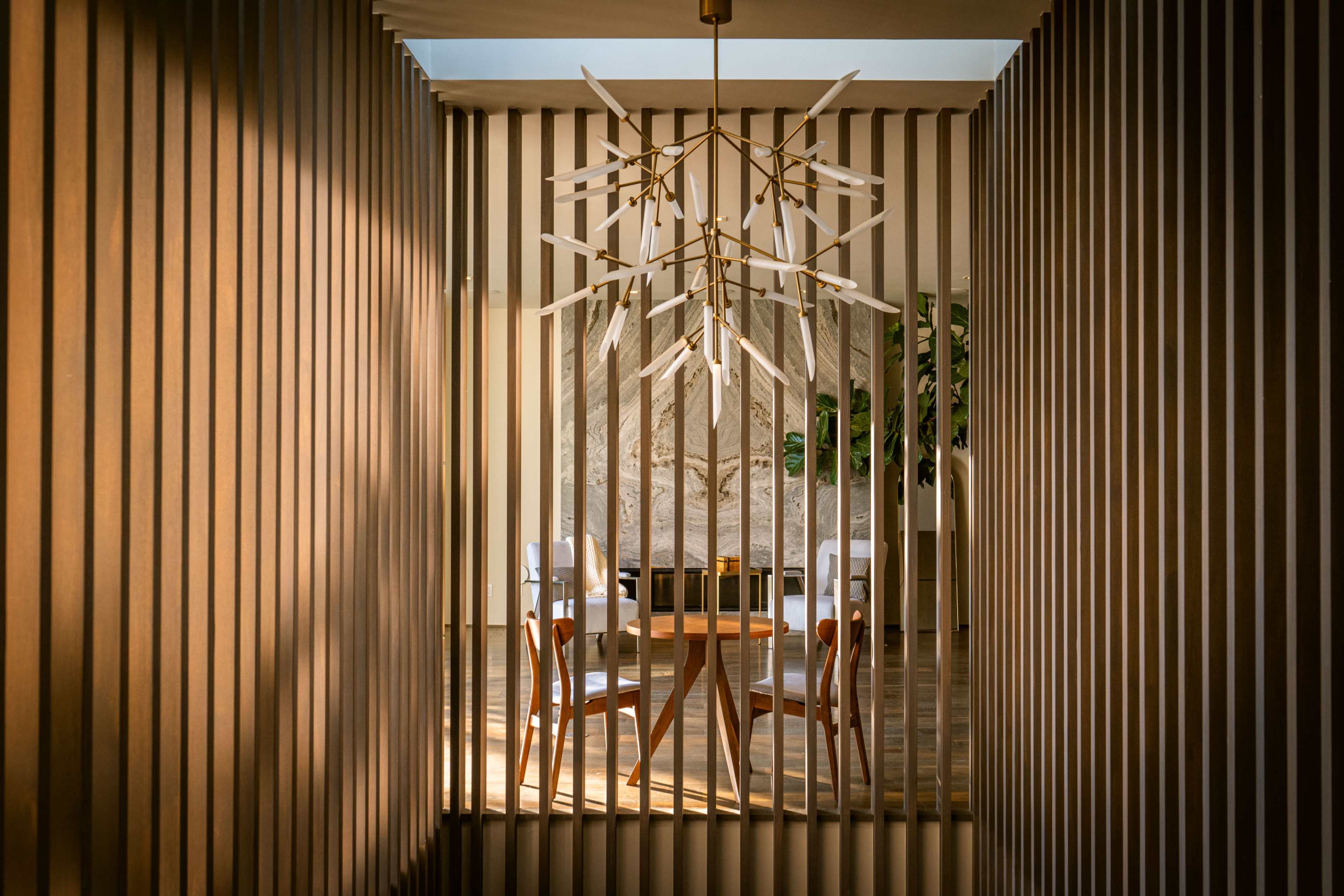 A modern interior space features a wooden slatted partition, a round table with chairs, and an intricate light fixture.